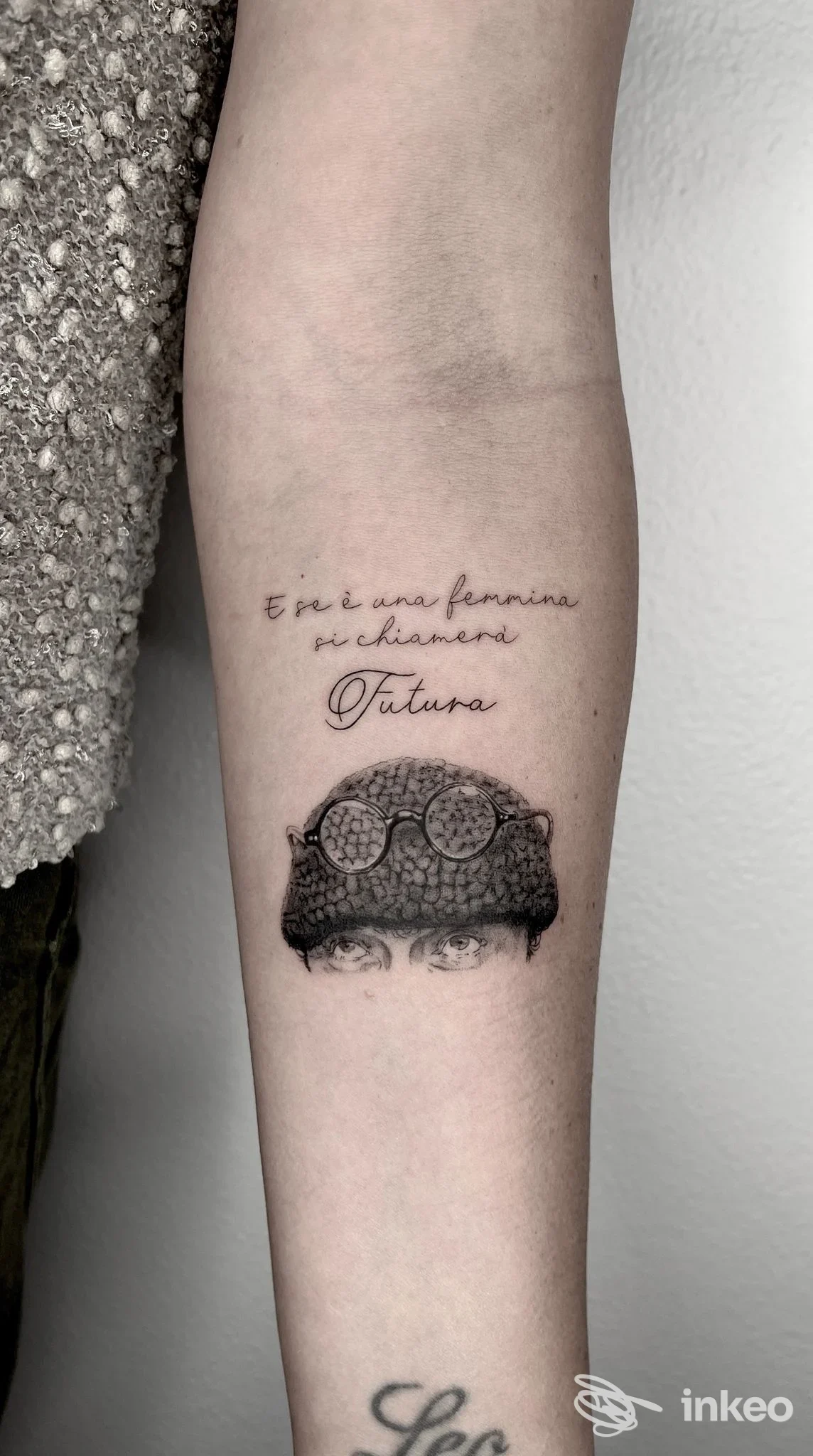 Tatuaggio sull’avambraccio: grafica scura con elementi geometrici o decorativi; testo in corsivo sopra la grafica.