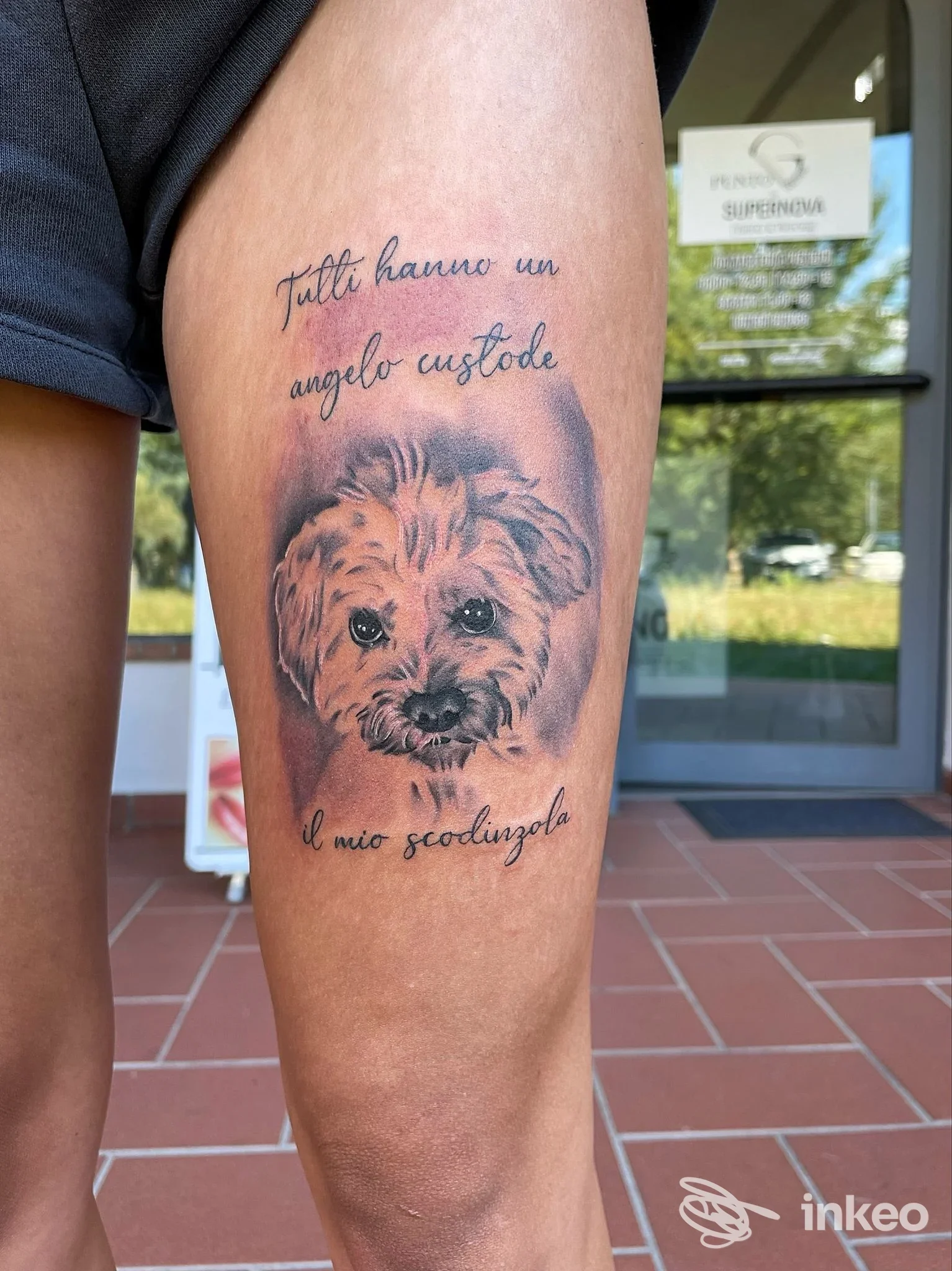Tattoo realistico di un cane su coscia, accompagnato da testo in corsivo italiano. foto scattata all'aperto vicino a uno studio. Il soggetto è direttamente visibile e il tatuaggio occupa la parte superiore della coscia.