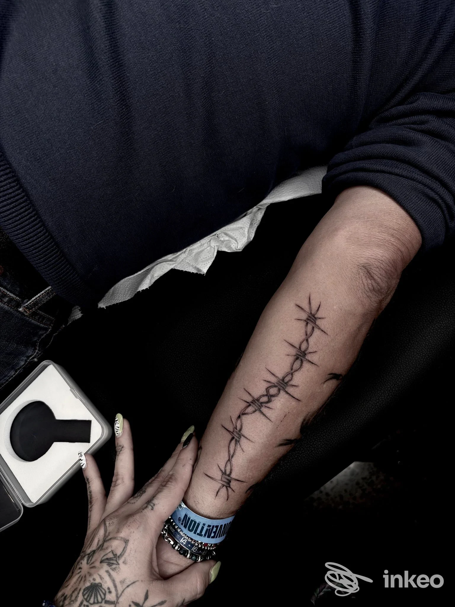 Tattoo sul avambraccio raffigurante una catena di filo spinato realizzata sulla pelle, postura della mano con anelli visibile.