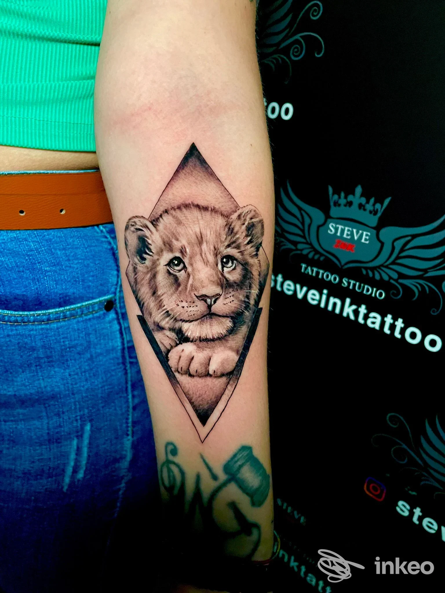 Tatuaggio realistico di un cucciolo di leone incorniciato in una forma a rombo sull’avambraccio sinistro, sfondo scuro, visibile solo la parte di braccio; studio Steve Ink Tattoo.