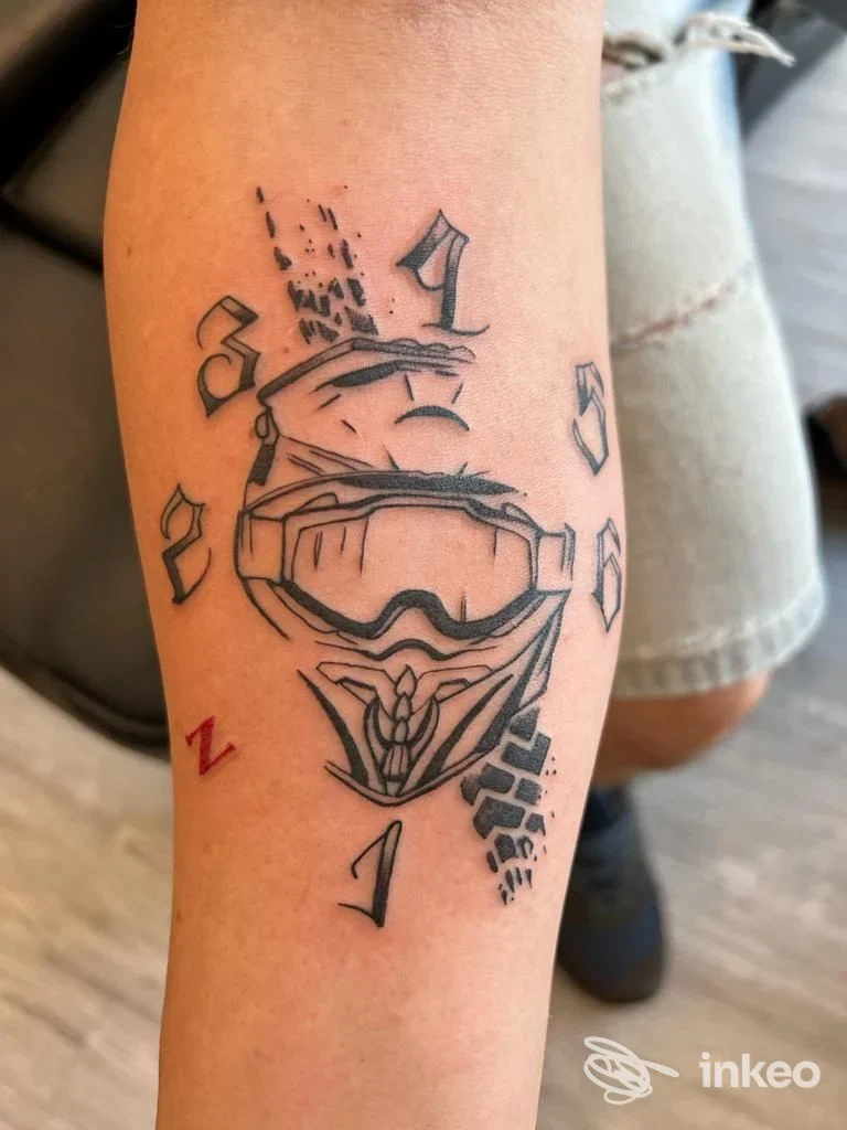 Tattoo visibile su avambraccio raffigurante una maschera da moto-cross con dettagli cromati, linee nette e piccoli elementi decorativi intorno.