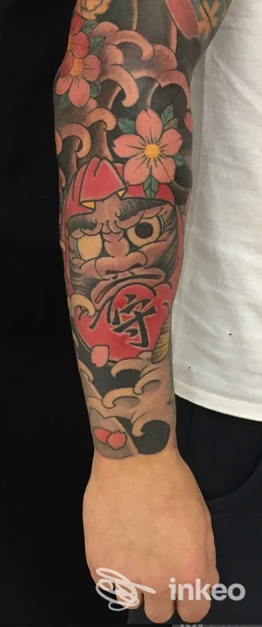 Avambraccio con una sleeve di tattoo colorati: fiori rosa, arancione e giallo con sfondo scuro, e un personaggio cartoonesco rosso al centro, realizzato sulla pelle.