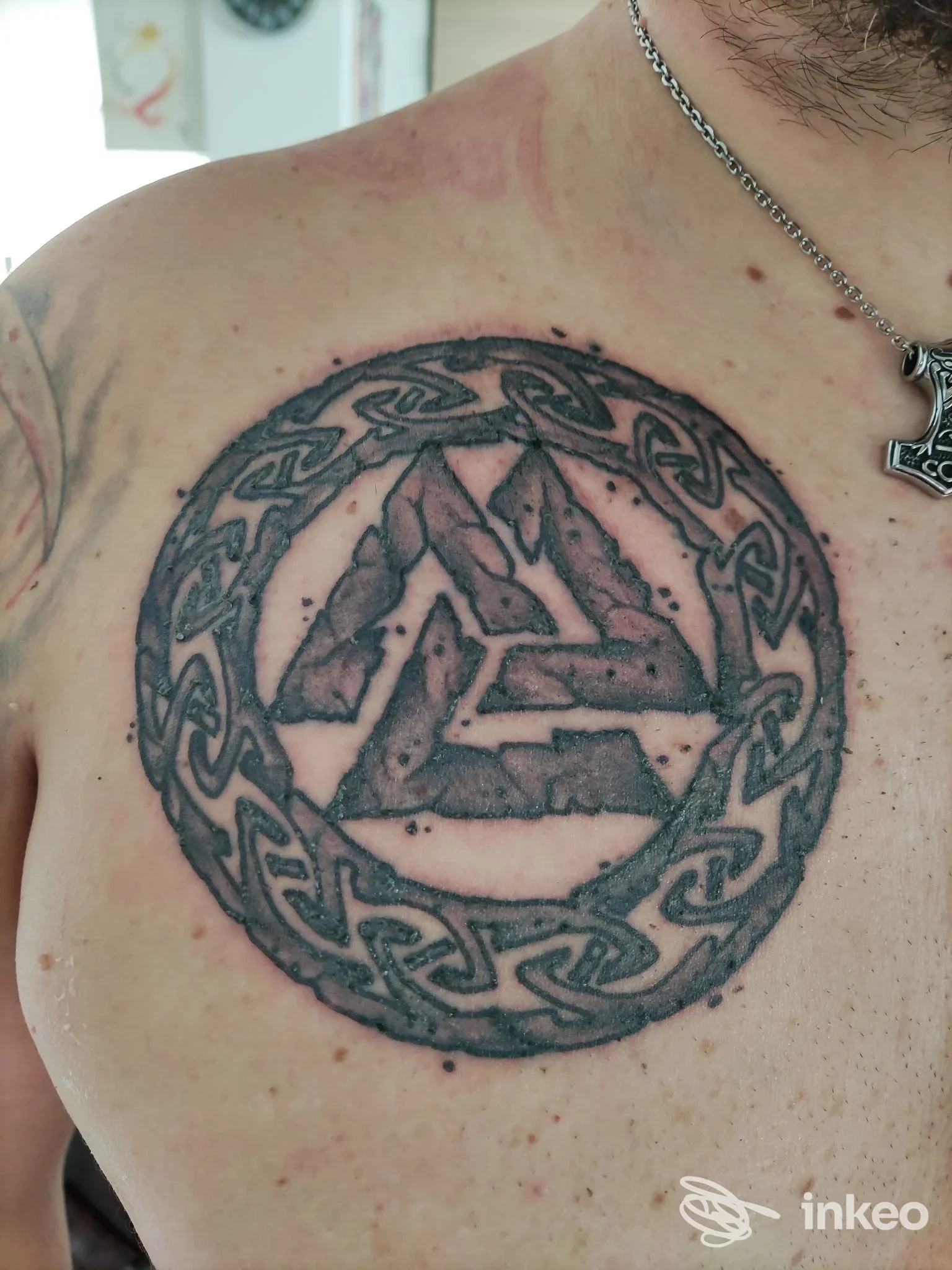 Tattoo circolare con motivo intrecciato celtico su petto sinistro, contenente triangolo centrale, contorno scuro e sfumature nere sulla pelle del soggetto.