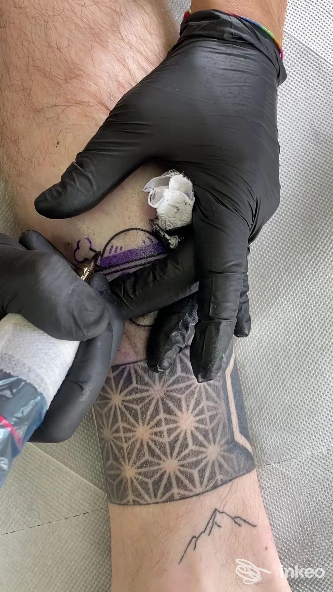 Inquadratura ravvicinata di una gamba con tatuaggio geometrico in corso; mani guantate lavorano sul disegno mentre sono presenti strumenti da tatuaggio e materiali.