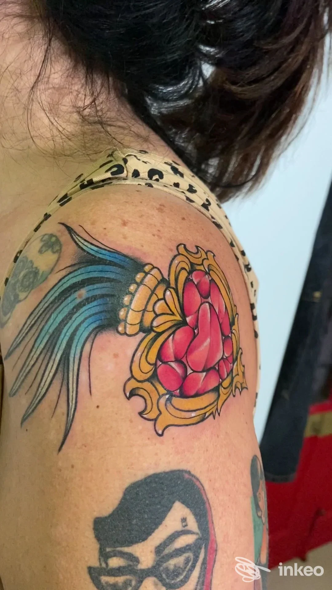 Tattoo colorato sulla spalla destra, con ornamento floreale azzurro e rosa, contorni neri ben definiti, visibile una seconda grafica in toni scuri. È una foto ravvicinata di una realizzazione corporea sulla pelle.