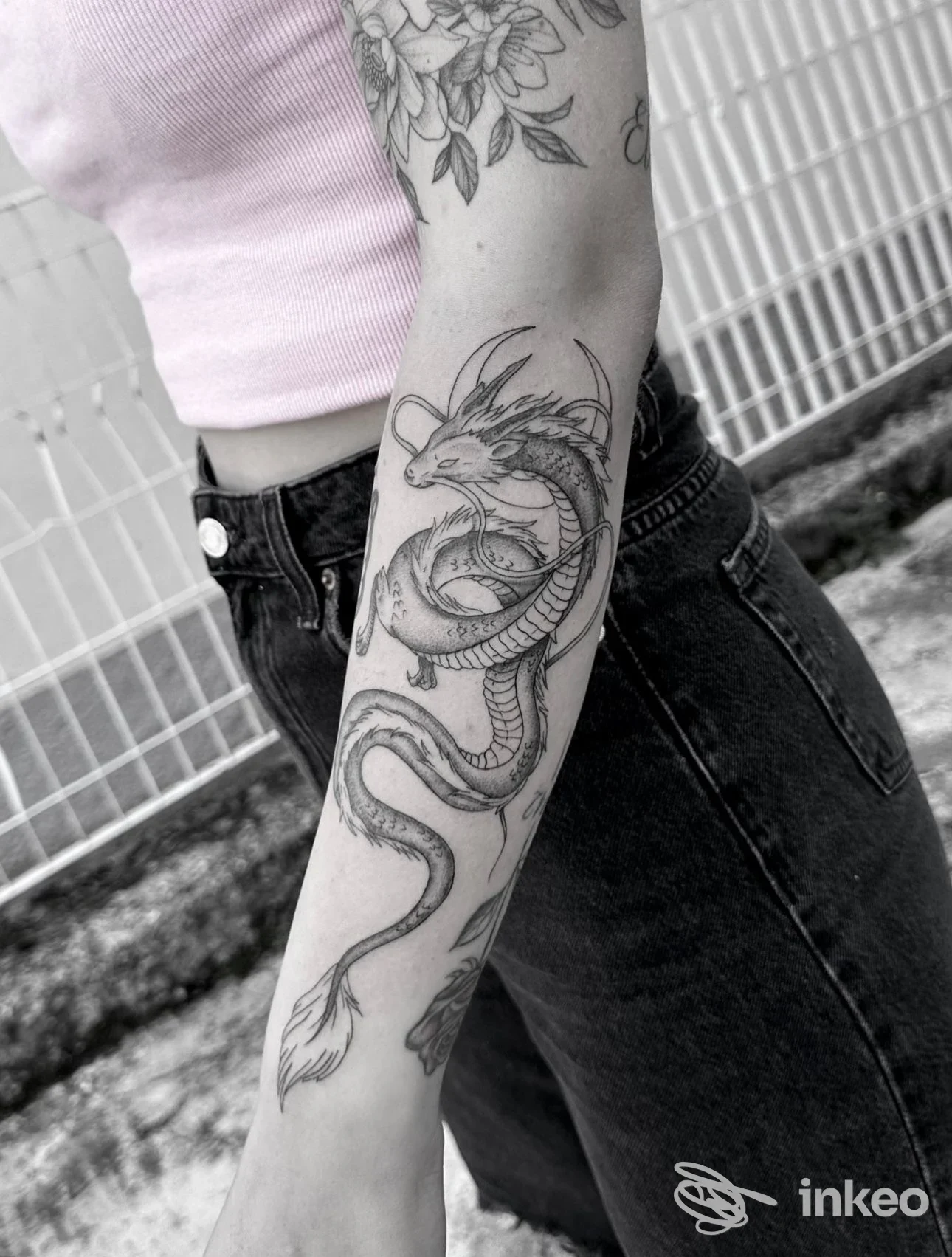 Tattoo su avambraccio raffigurante drago serpentino, disegno in bianco e nero su pelle, visto con maglietta rosa e jeans scuri, sfondo con ringhiera.