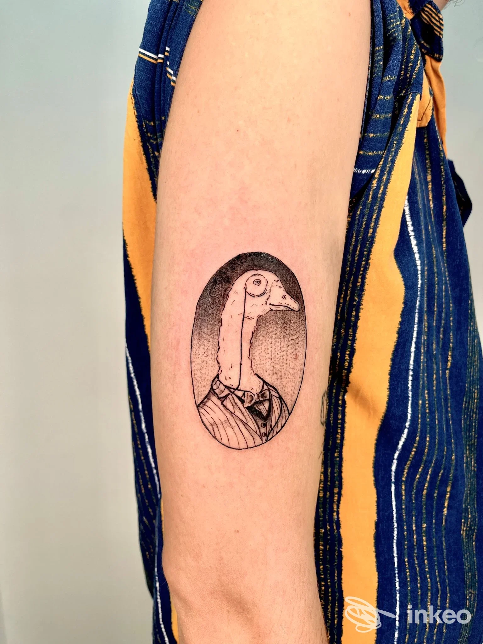 Tatuaggio realizzato sull’avambraccio: un uccello antropomorfo in abito, racchiuso in un ovale, eseguito in stile line art. La pelle è nuda, senza contenuti promozionali o alterazioni visive.
