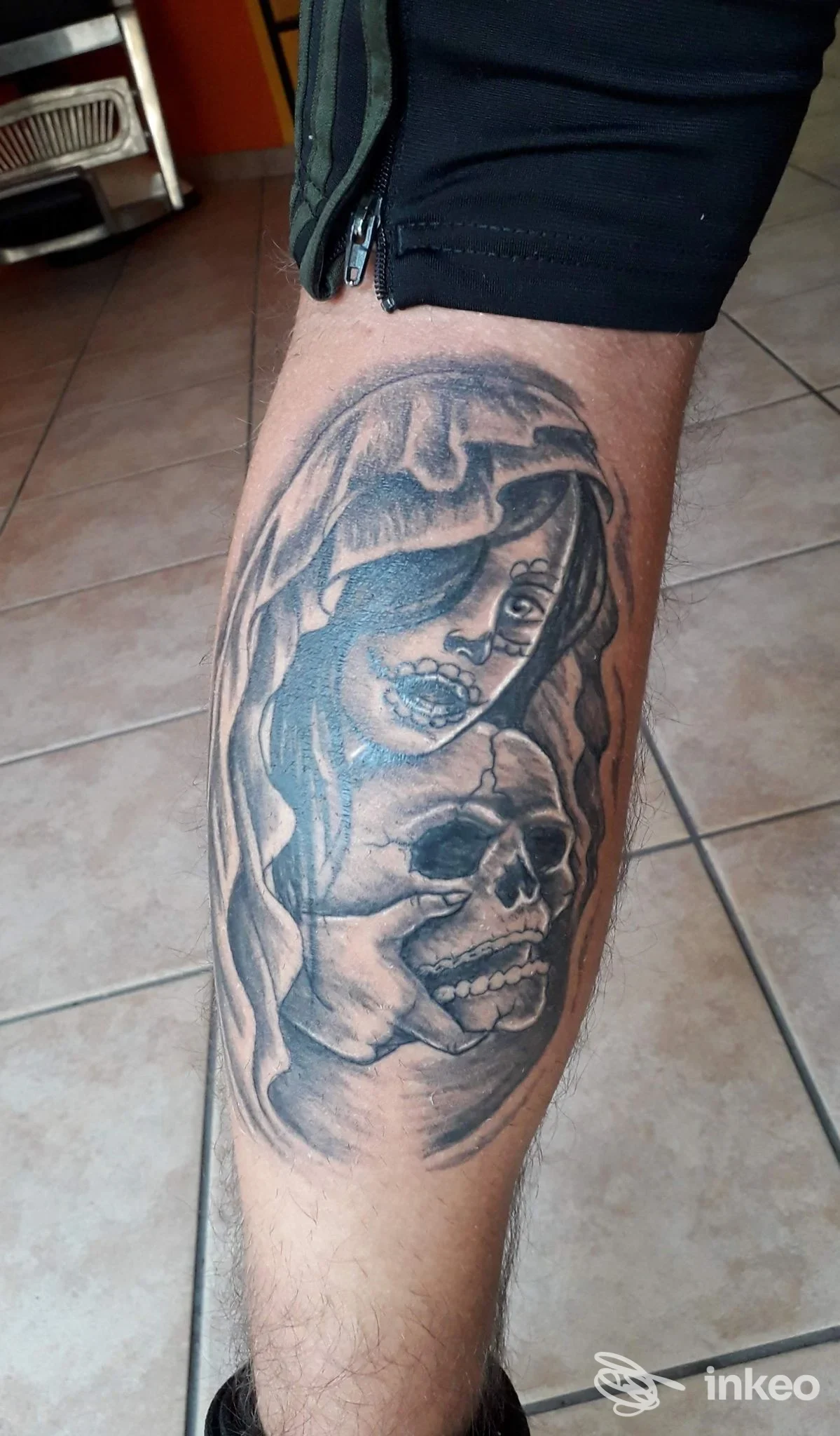 Tatuaggio realistico in bianco e nero su polpaccio raffigurante una figura femminile avvolta da un mantello accanto a un teschio, stile realistico, materiale corporeo su pelle nera.