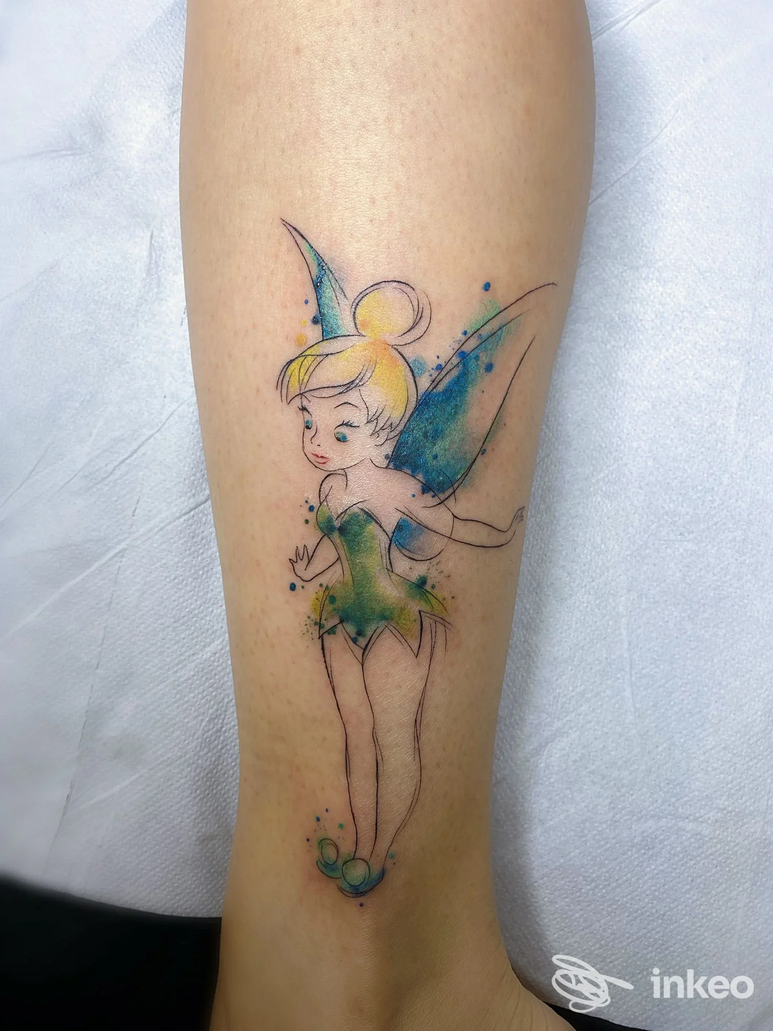 Tatuaggio su pelle raffigurante una fatina colorata con vestito verde e ali azzurre, realizzato con stile acquerello sulla gamba.