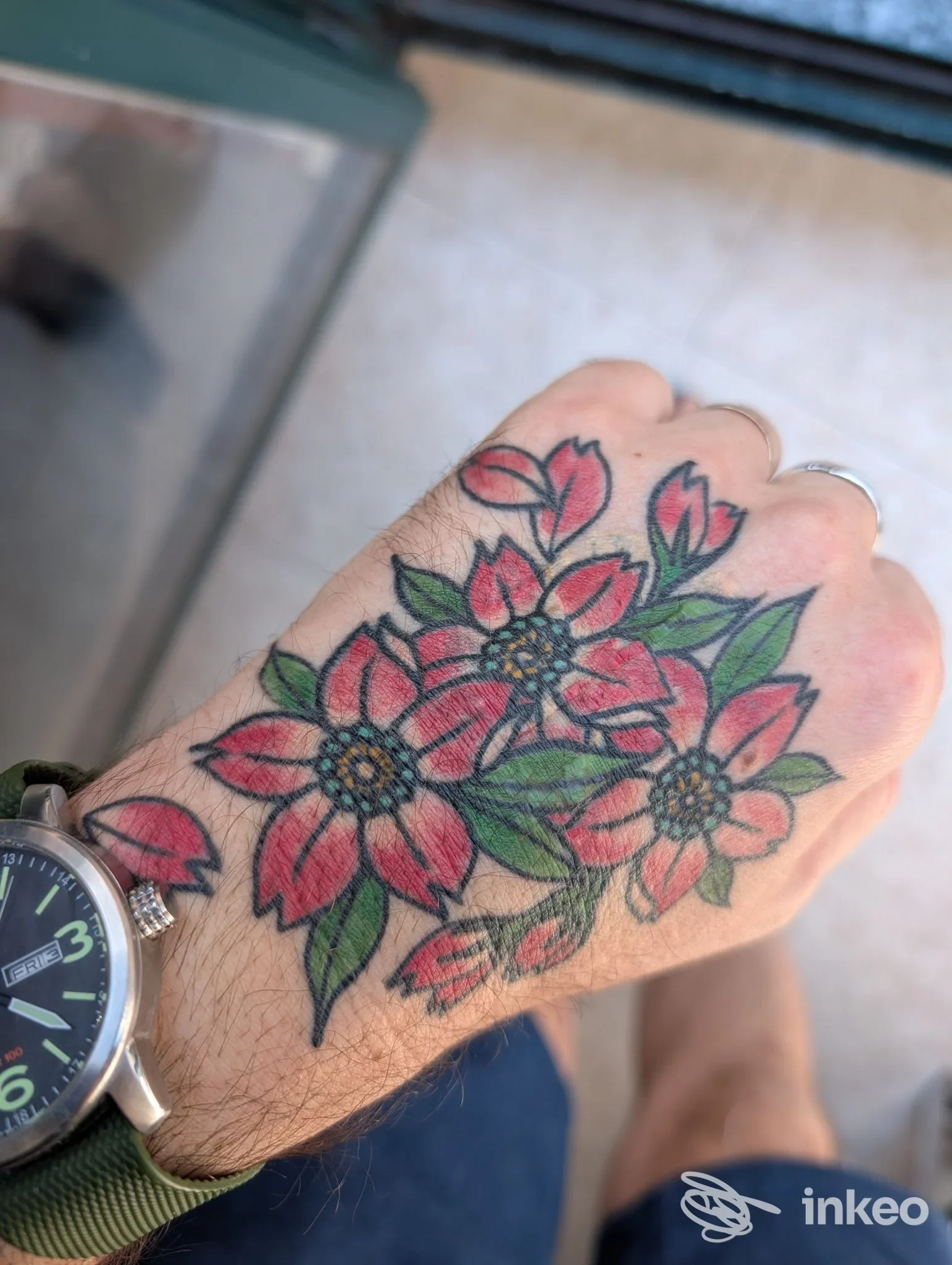 Tattoo floreale colorato visibile sulla mano, bordi neri definiti e sfumature rosse e verdi; eseguito su pelle presso Freak Tattoo Shop, Como.
