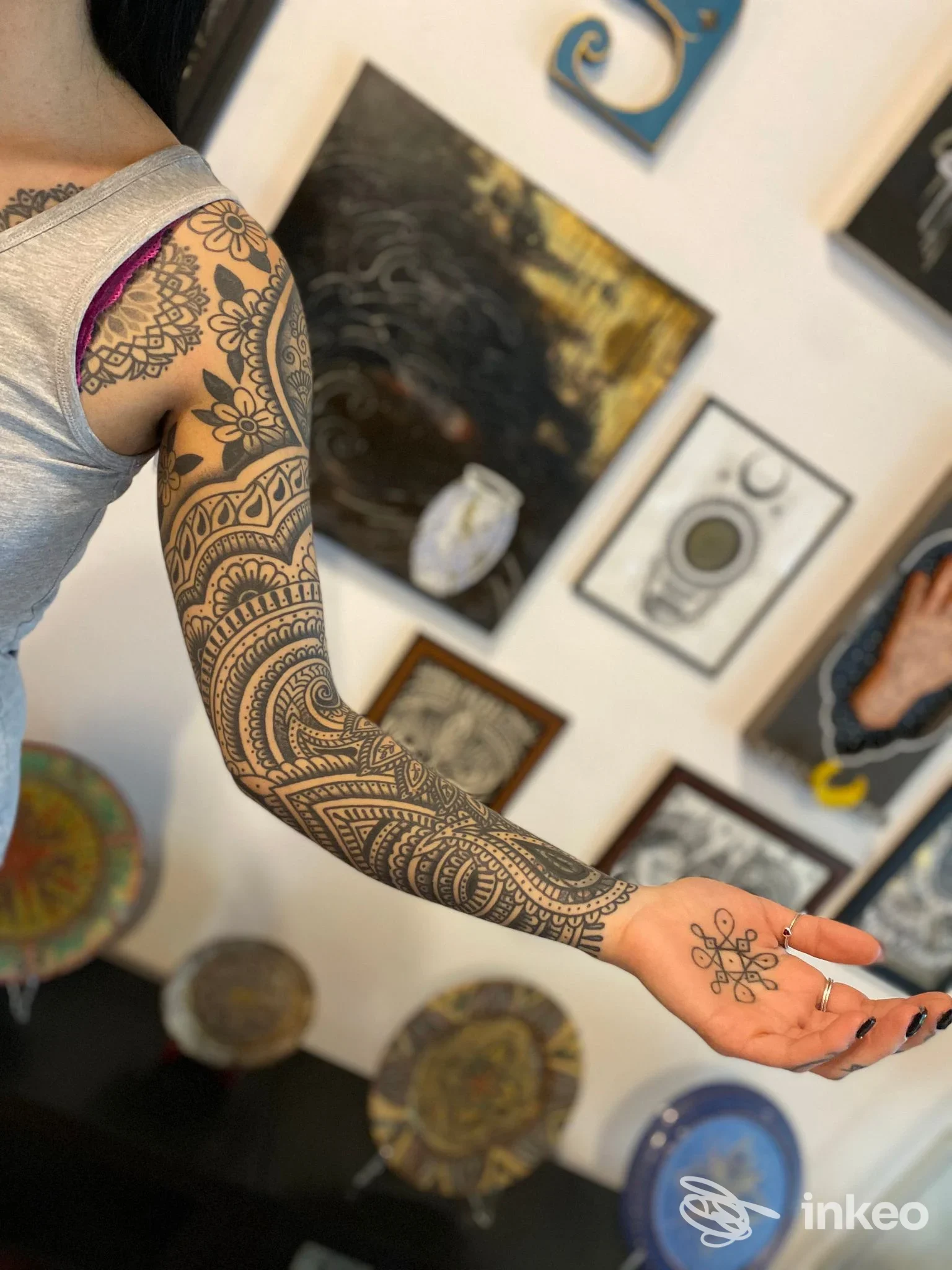 Braccio sinistro con tatuaggio a manica in stile geometrico, disegnato sulla pelle reale, visibile in un ambiente di studio con opere d'arte alle pareti.