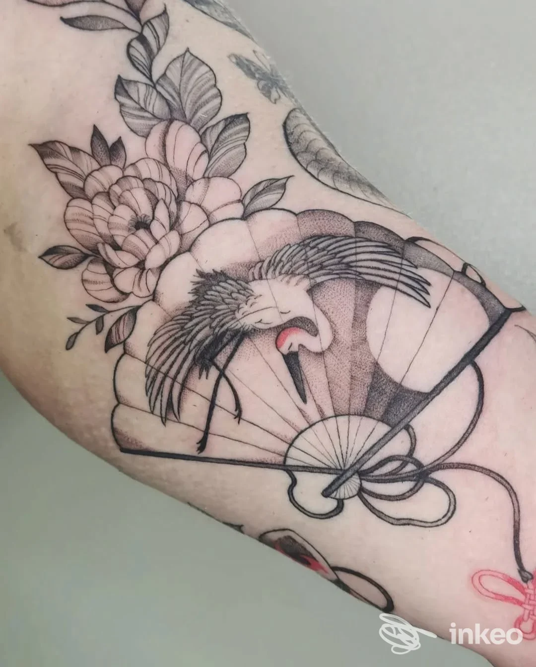 Tatuaggio sull'avambraccio raffigurante una gru con fiori e ventaglio, linework con sfumature leggere, nessun volto visibile.
