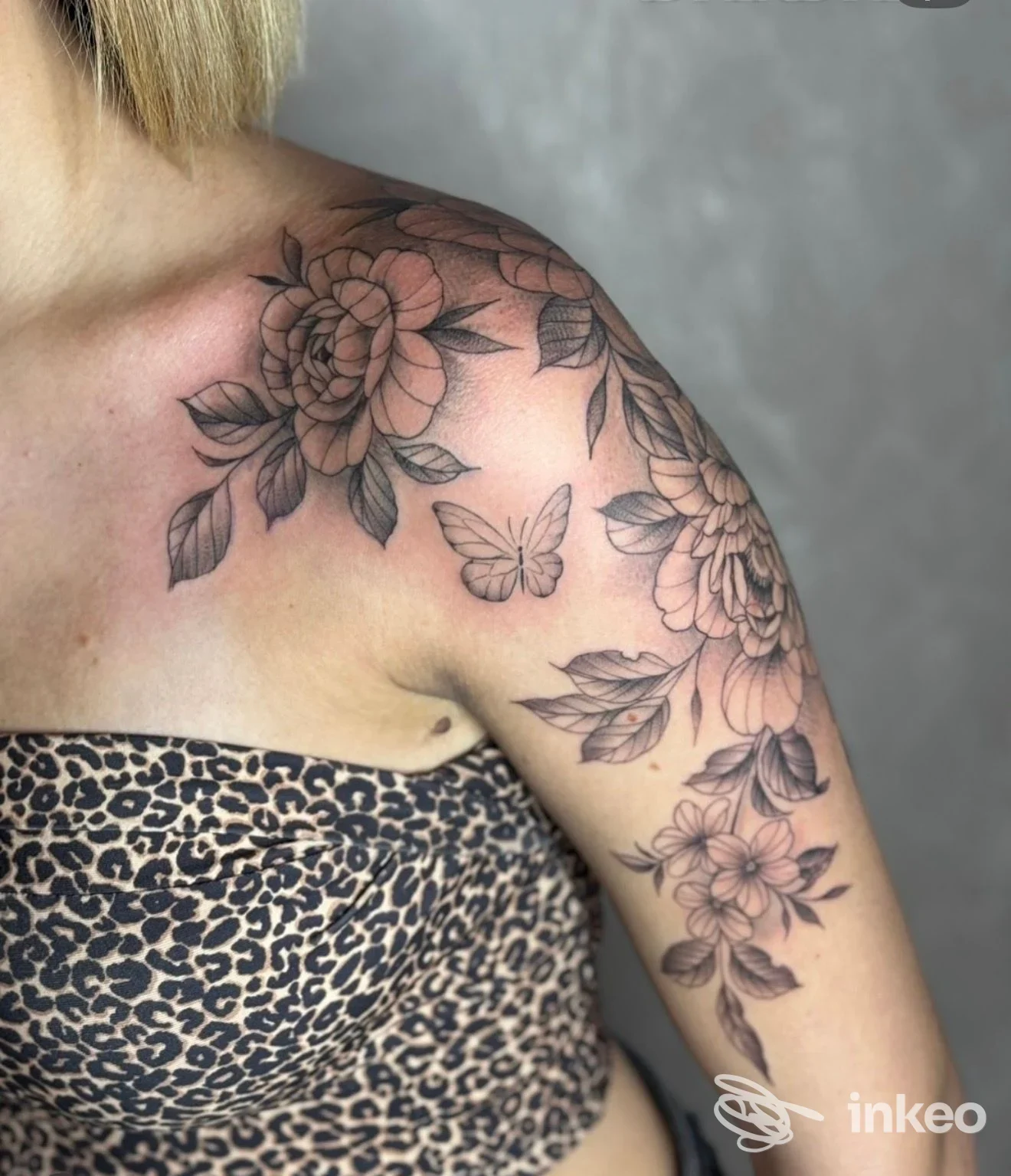 Tattoo floreale in linea nera su spalla e braccio, con fiori e una farfalla, su pelle reale senza volto visibile.