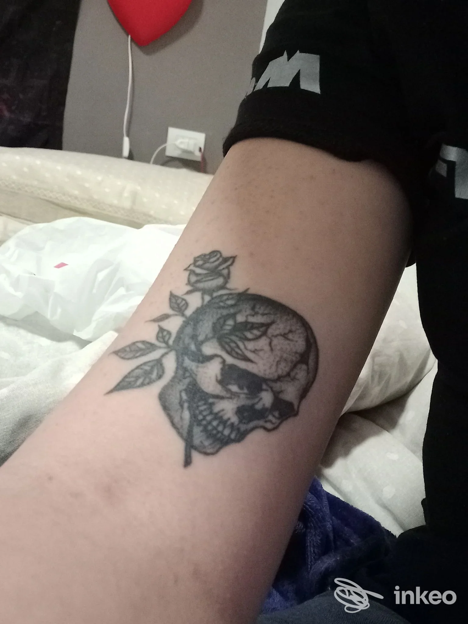 Tatuaggio in bianco e nero su avambraccio raffigurante una testa di teschio circondata da rami e fiori, su pelle reale.