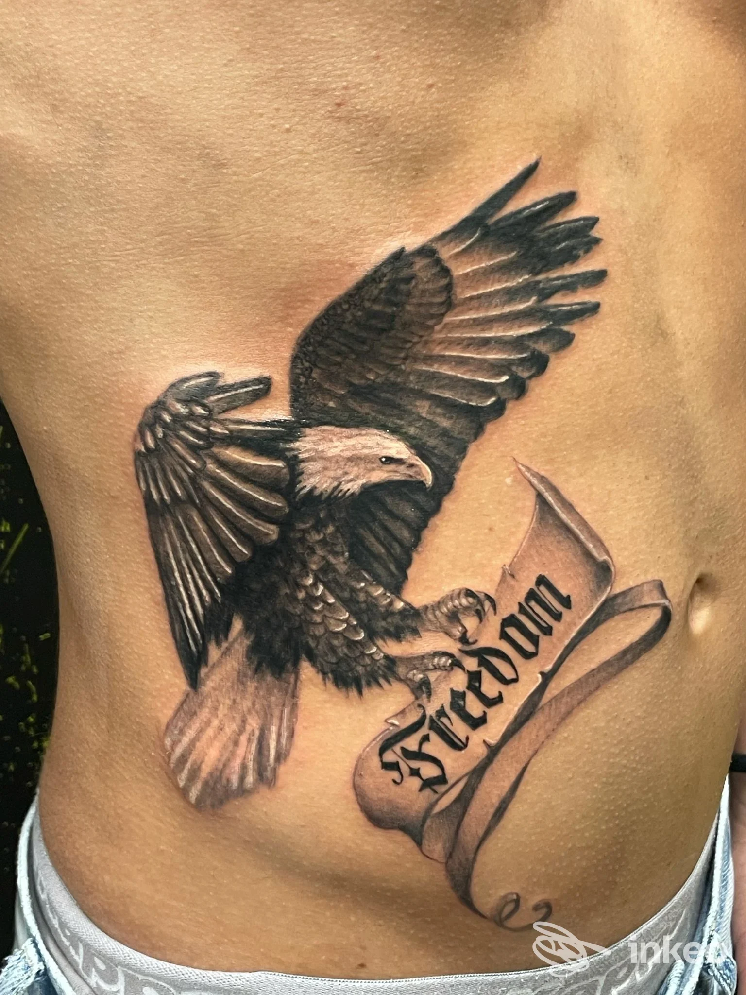 Tattoo realistico di un corvo in volo su fianco sinistro del corpo, accompagnato da una fascia con testo elegante. LUOGO non specificato.