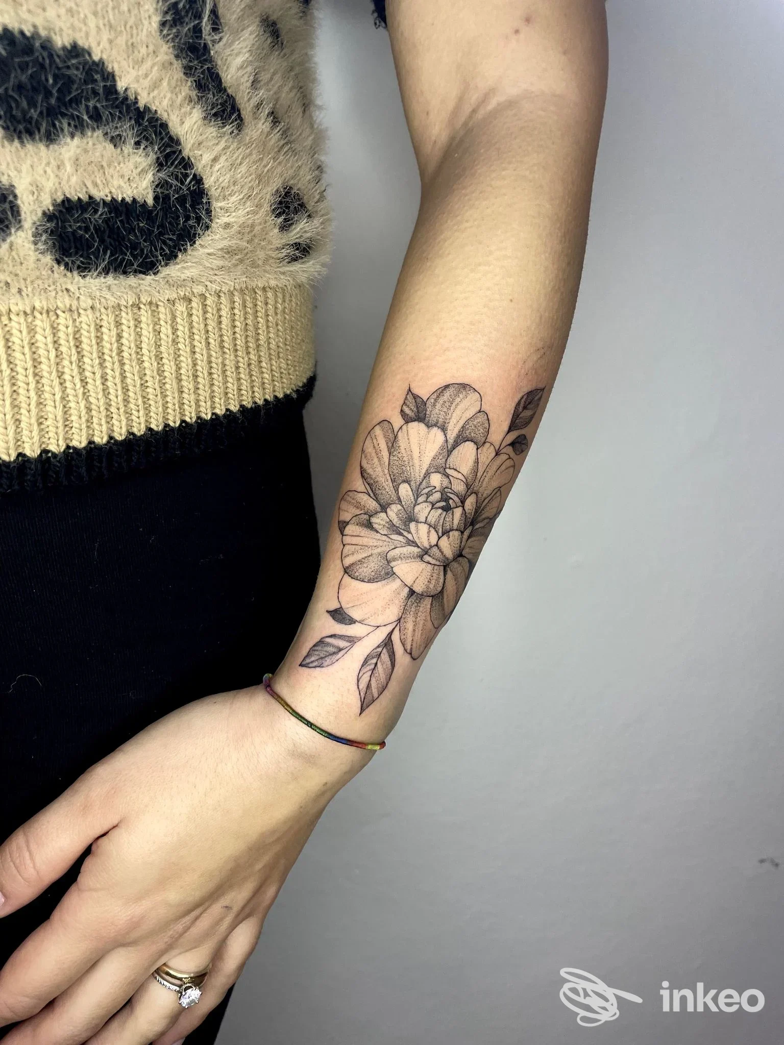 Tattoo floreale nero/grigio sull'avambraccio, disegno di fiori e foglie; parte della maglia beige in cornice.