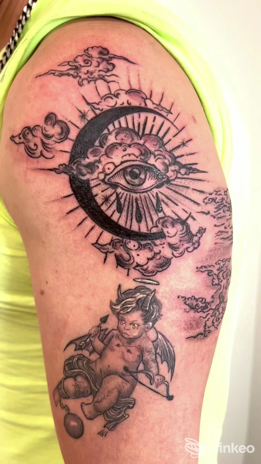 Tattoo visibile su braccio: luna con occhio circondata da nuvole, accompagnata da un cherubino alato; lavoro sulla pelle, nessun volto intero visibile, senza overlay sociale o immagine di schermo.