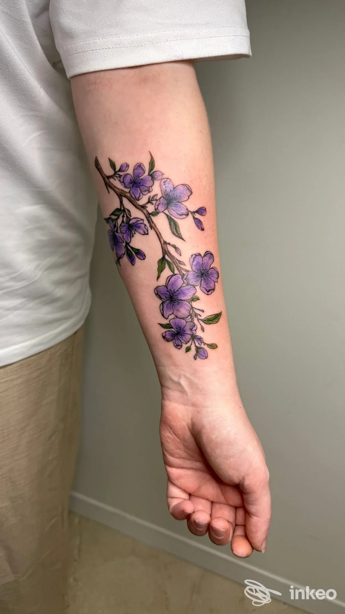 Avambraccio sinistro con tatto fiori viola lungo un ramo, realizzato sulla pelle; sfondo neutro, nessun volto visibile, grafica pulita e colori vividi.