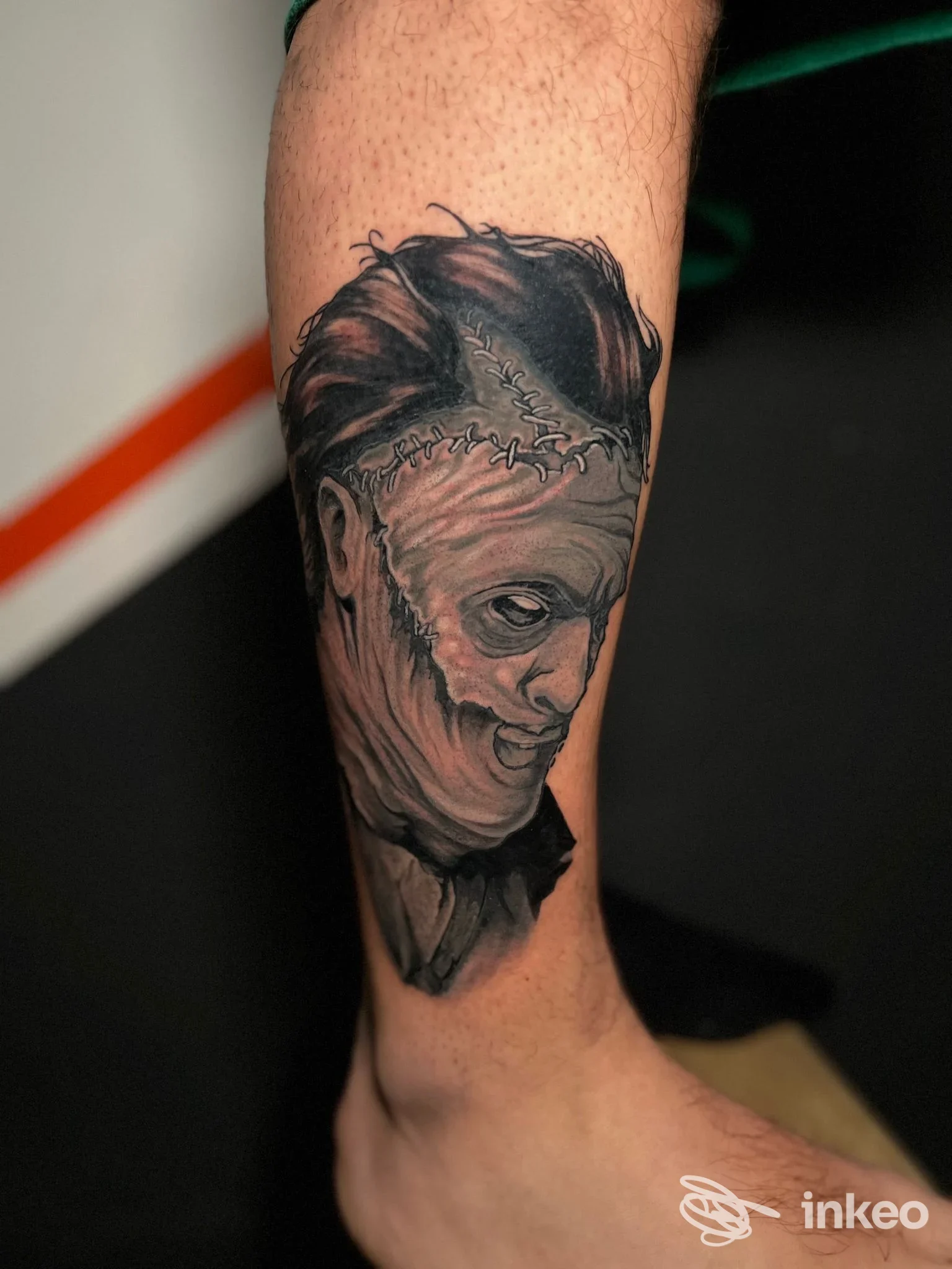 Tattoo realistico sul polpaccio raffigurante un volto stile mostruoso con cuciture sul cranio, tonalità in bianco e nero.