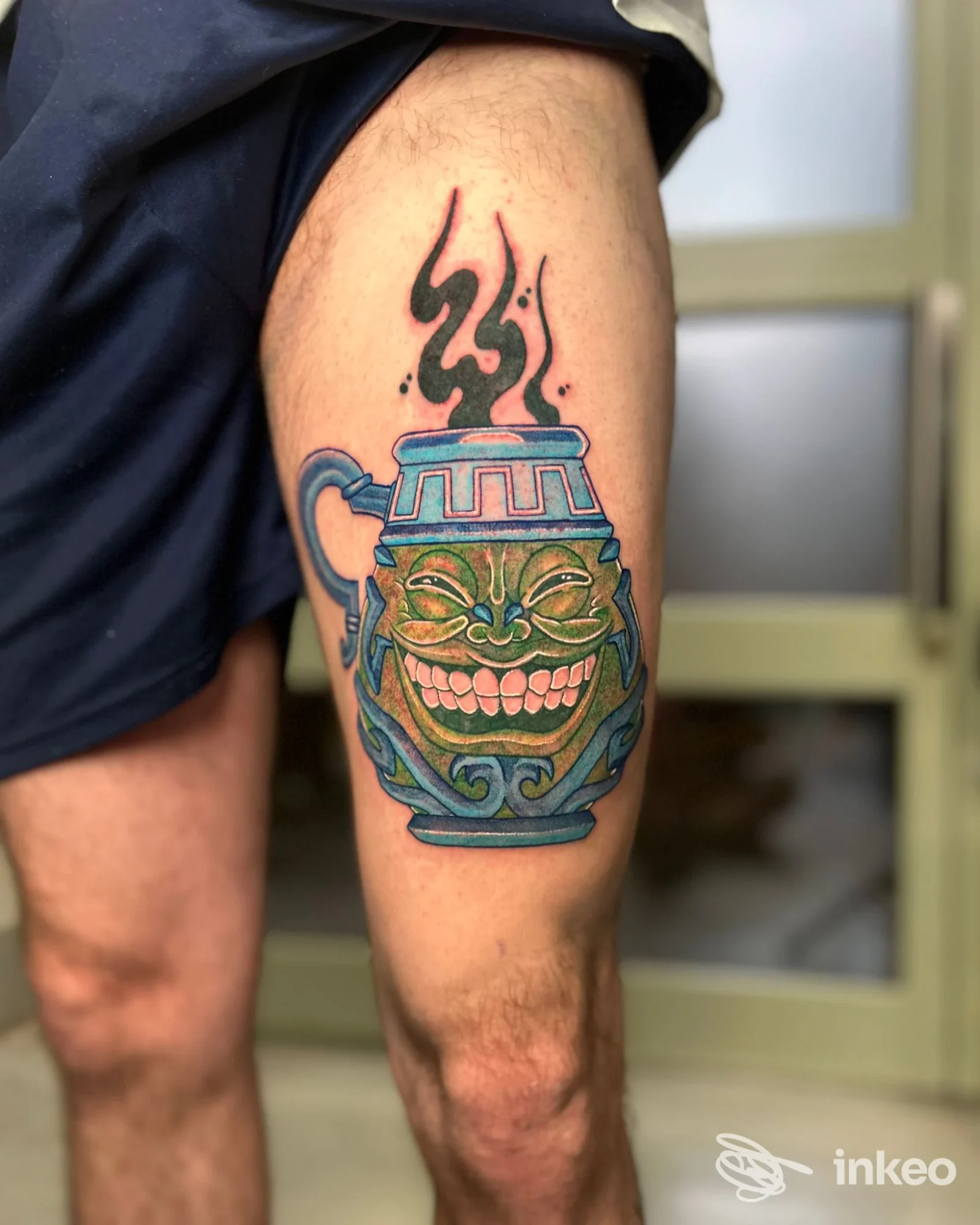 Tattoo colorato su pelle raffigurante una tazza decorata con una faccia sorridente, fumo nero che ne esce, applicato sulla gamba di una persona in posa laterale.
