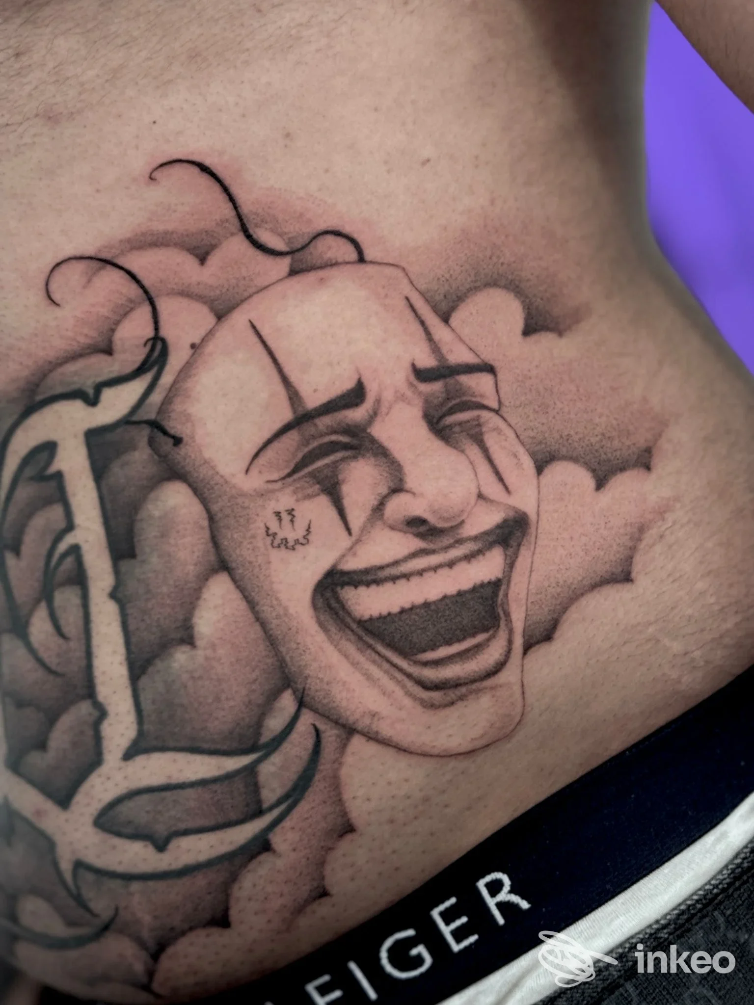 Tatuaggio su costola raffigurante una maschera teatrale sorridente tra nuvole, con tratto deciso e ombreggiatura.