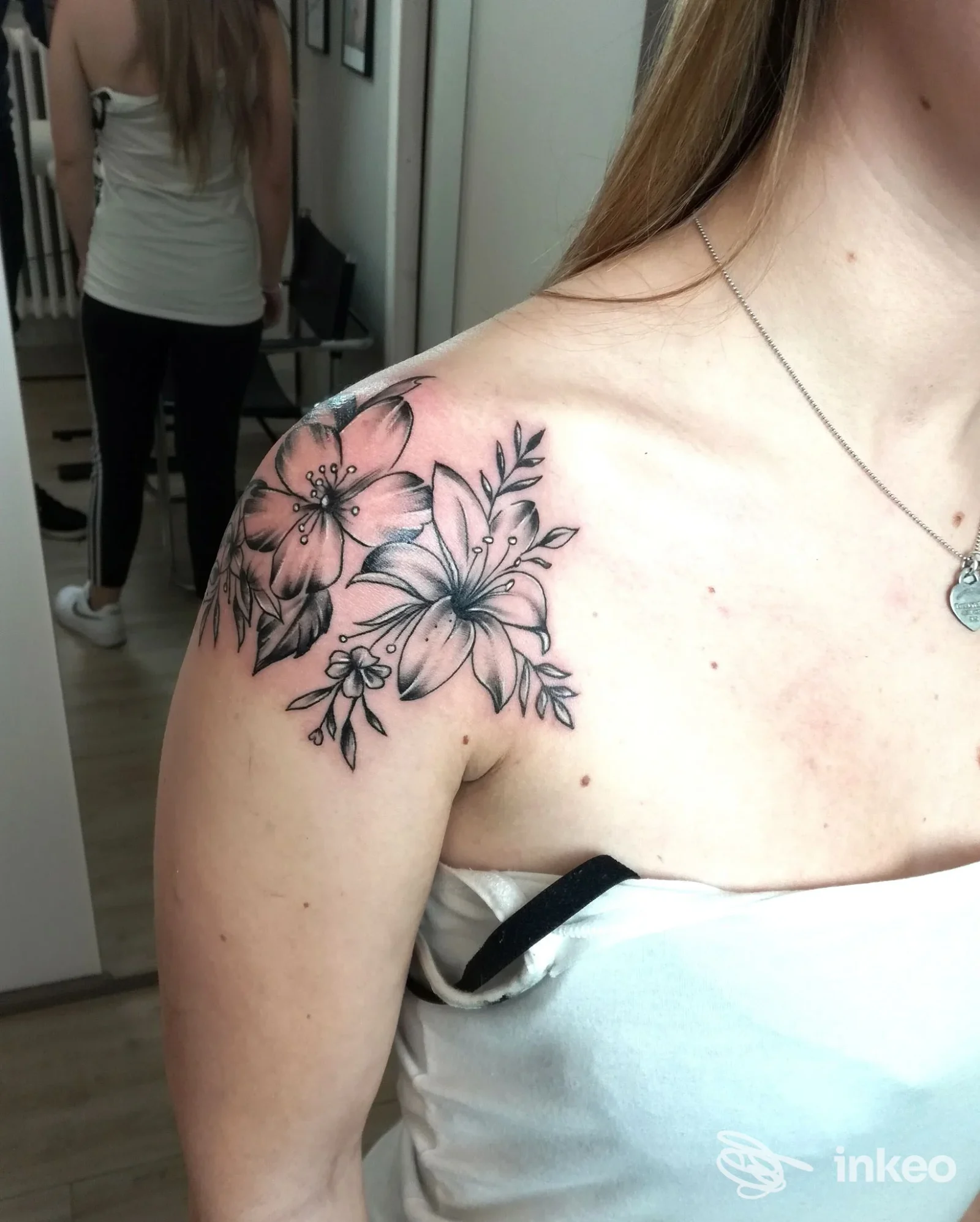 Tattoo floreale sulla spalla sinistra di una persona, disegno a linee nere con fiori e foglie.