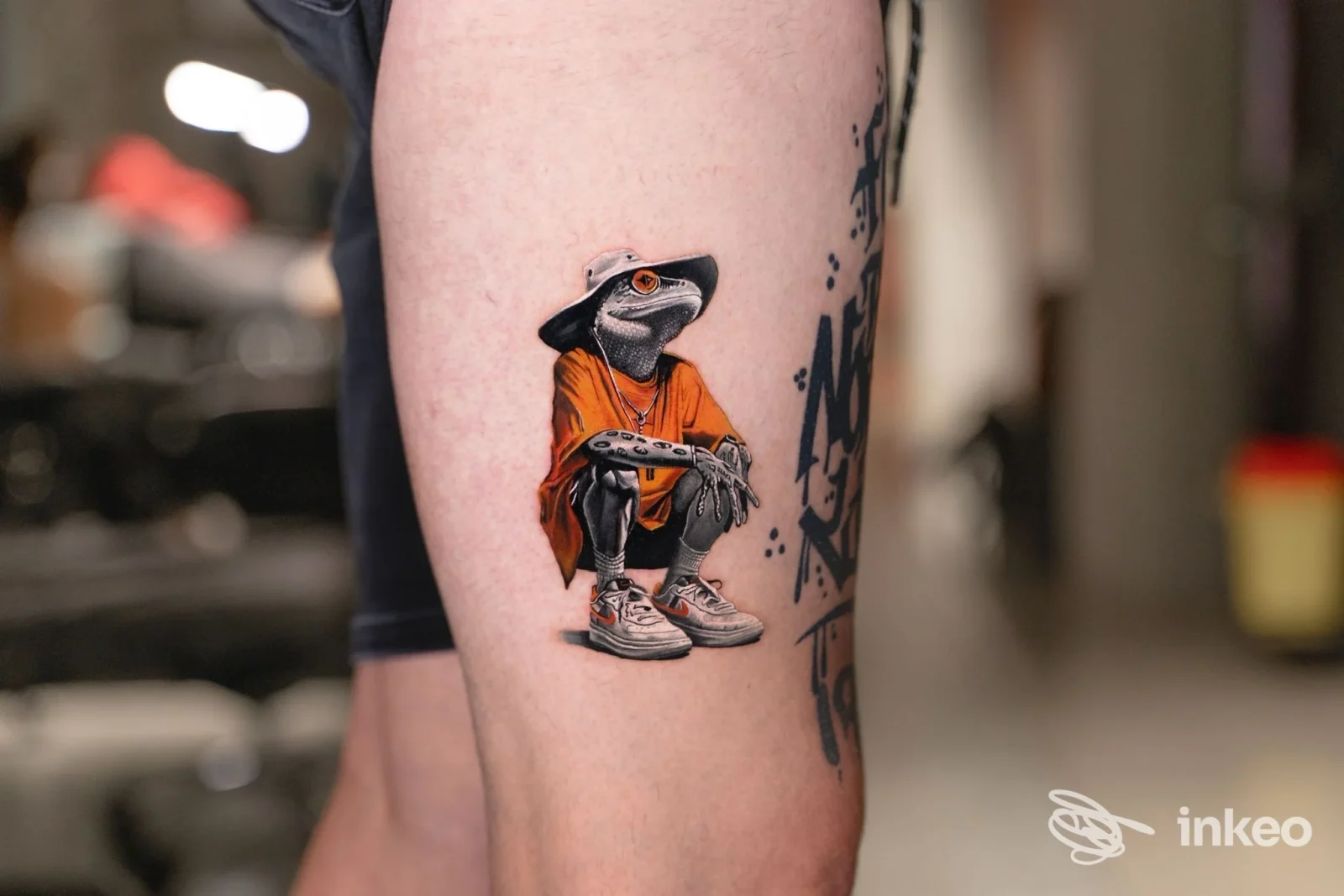 Tattoo raffigurante una figura antropomorfa con cappuccio arancione sul polpaccio di una persona, eseguito direttamente sulla pelle. Sfondo di studio sfumato.
