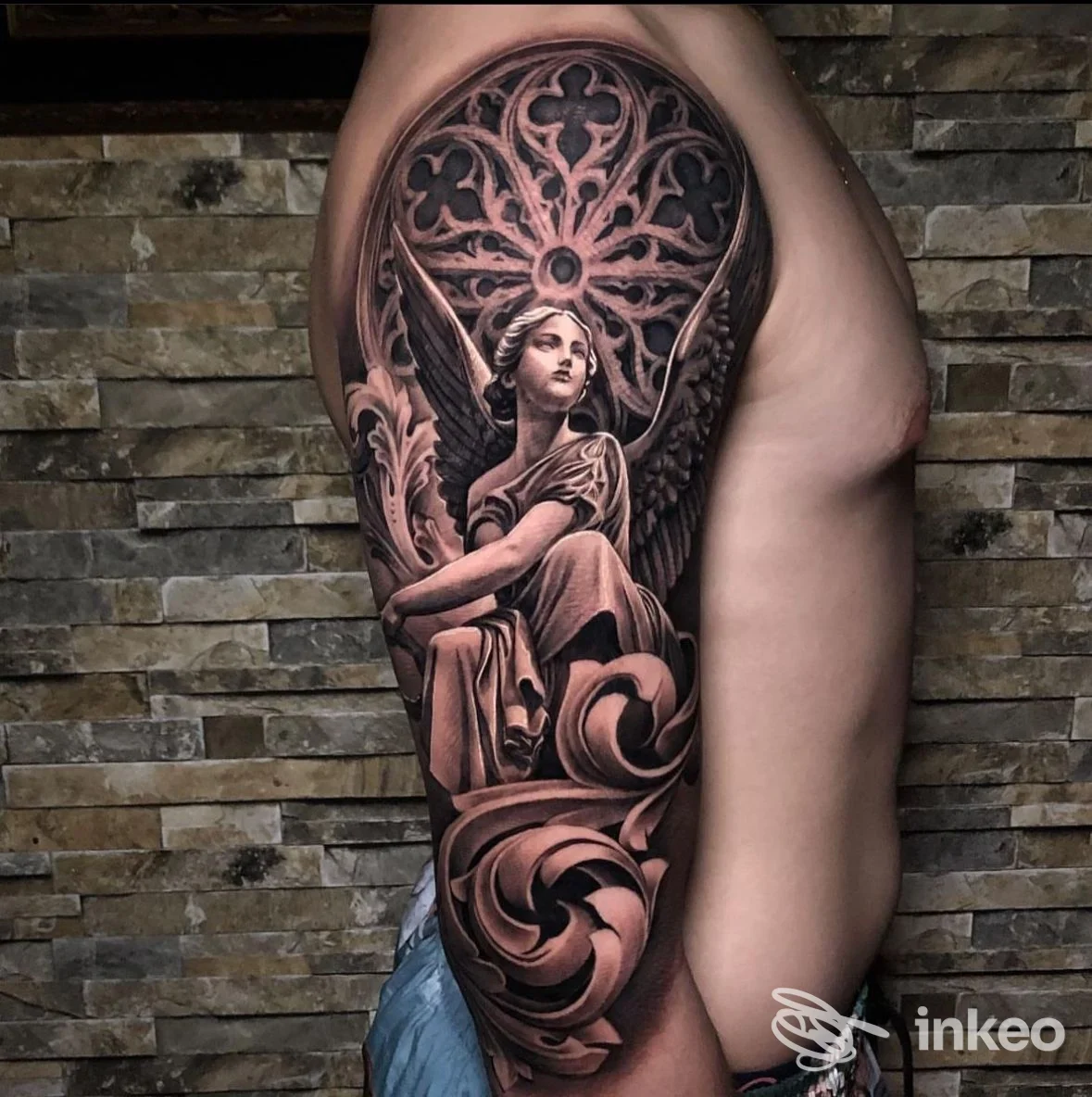 Tattoo realistico su pelle visibile sulla coscia: figura angelica e motivi decorativi in tonalità nero/grigio, sfondo in pietra, senza volto visibile.