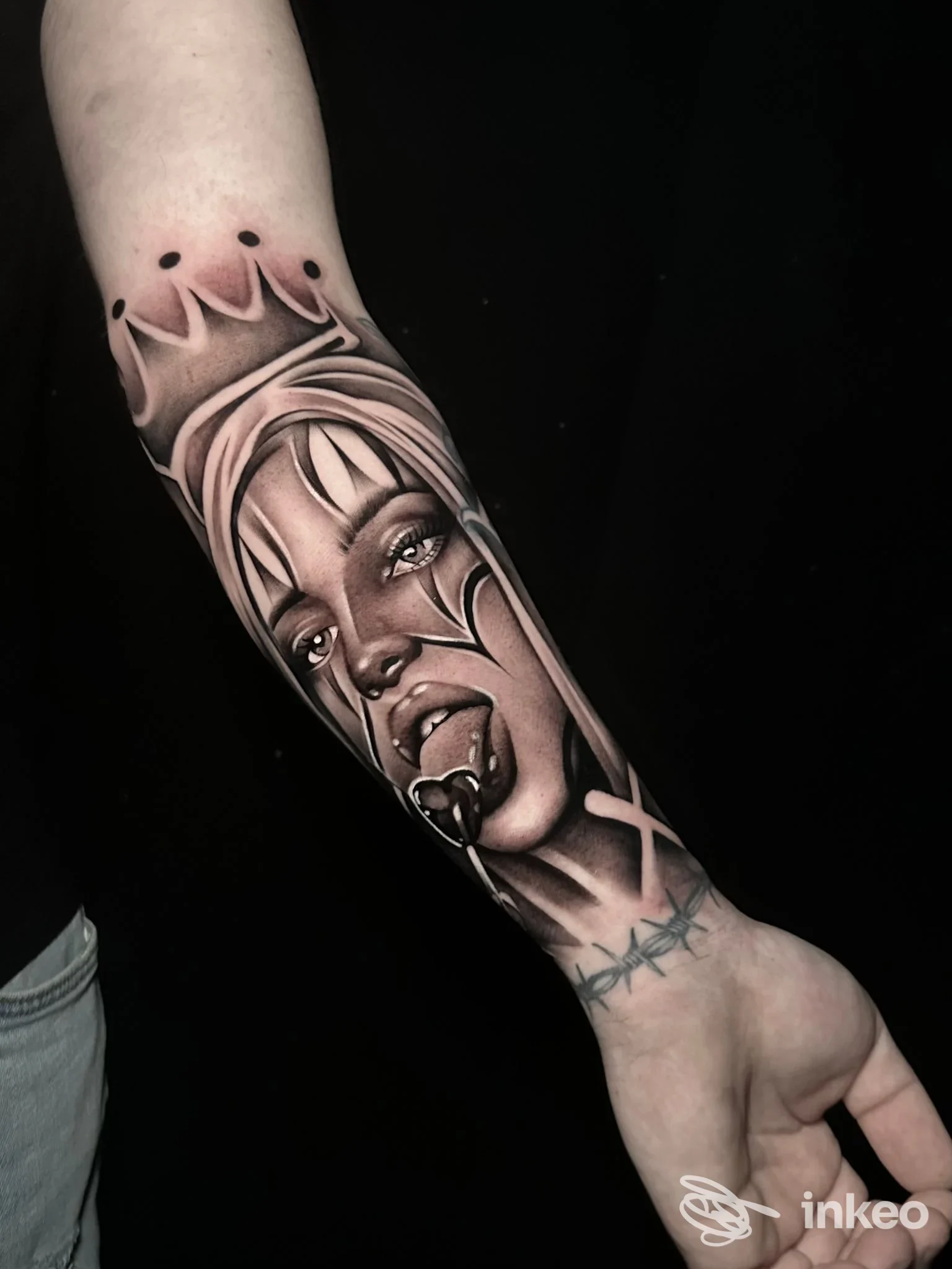 Tatuaggio realistico su avambraccio raffigurante volto femminile con corona e lingua fuori, realizzazione corporelle ben definita su pelle scura.