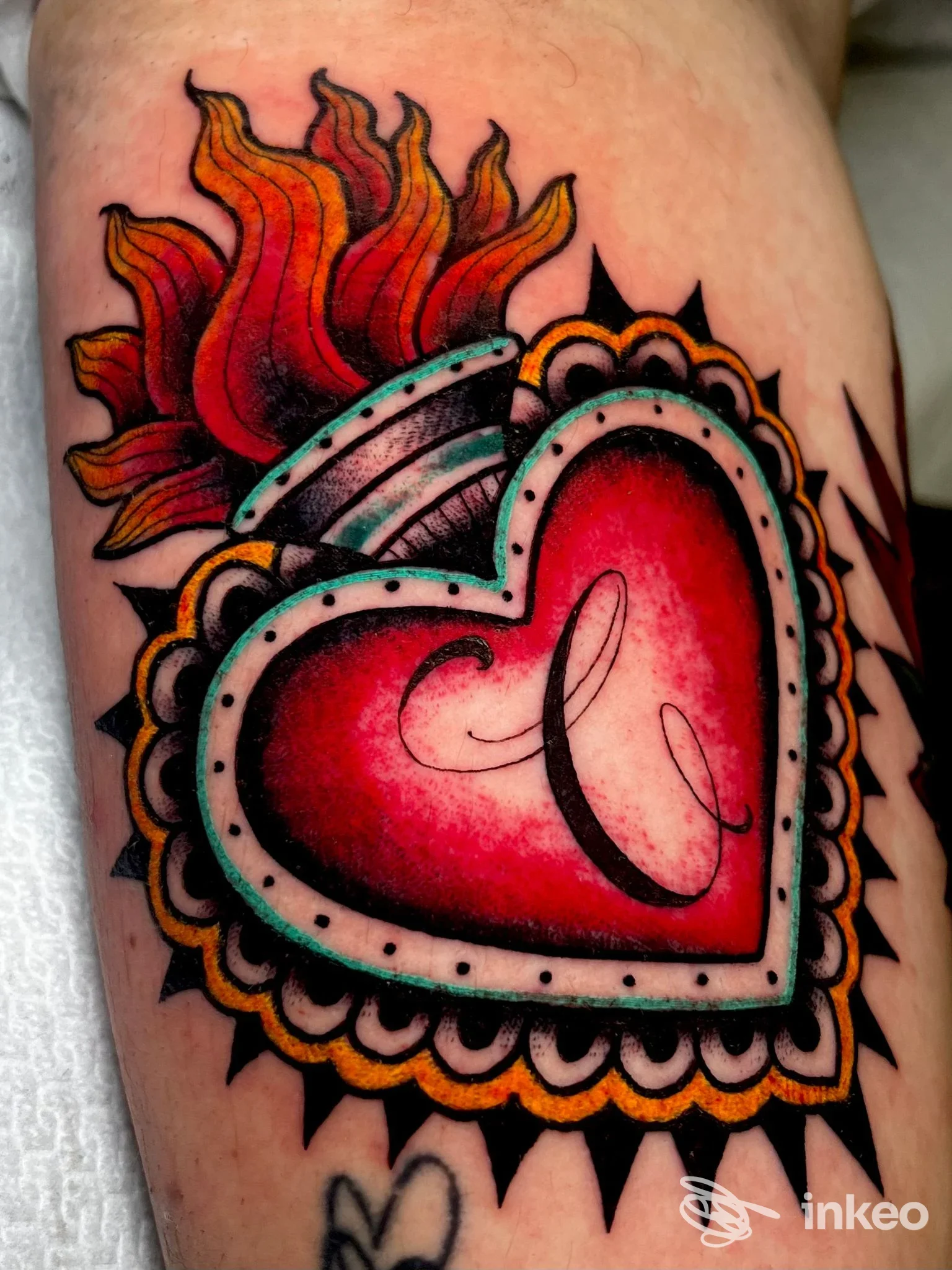 Tattoo a cuore rosso con contorni bianchi e ornamenti, fiamme arancioni alle sue spalle; tatuaggio sul corpo, visto in primo piano, firma dello studio House of Ink.