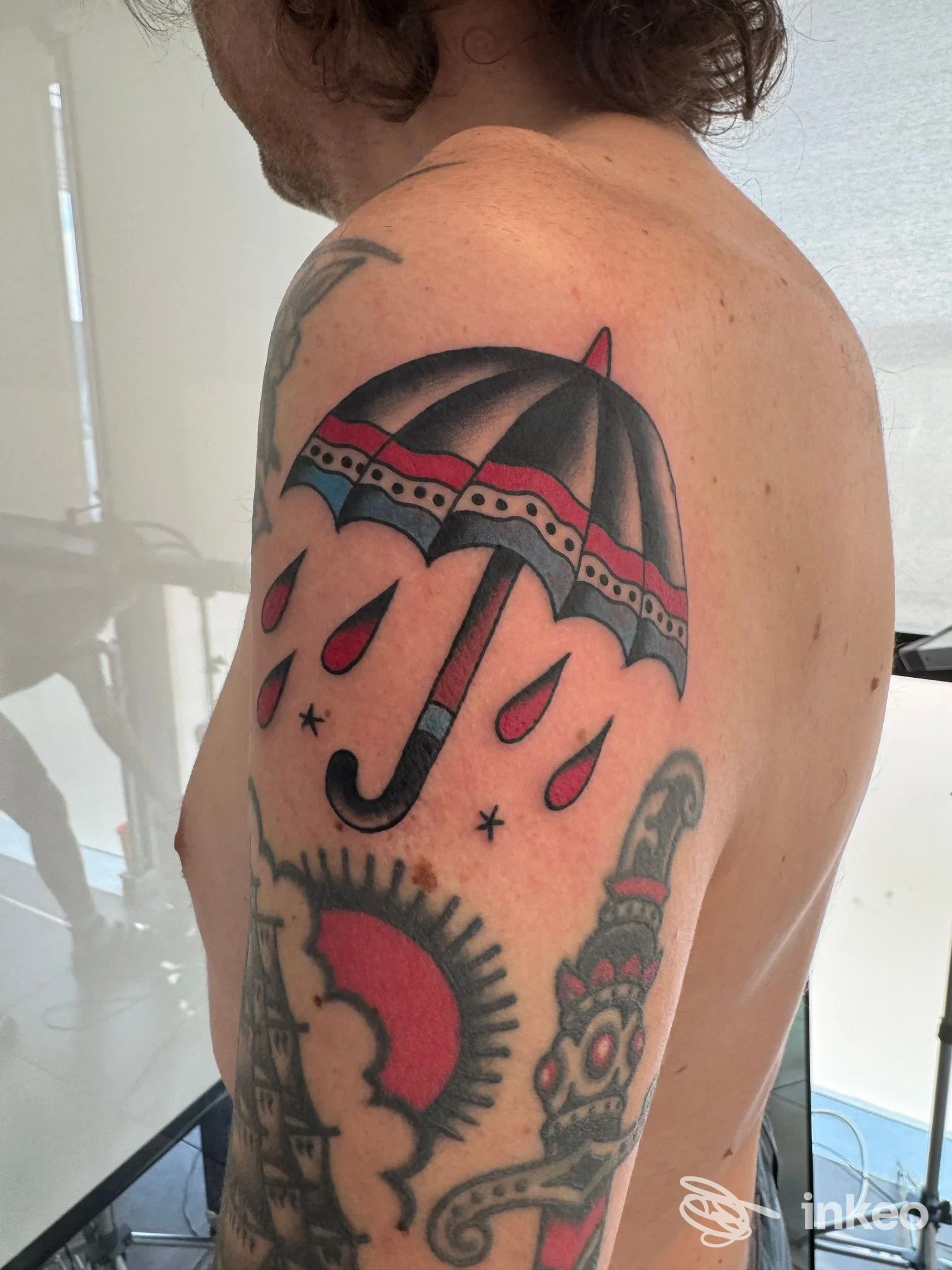Tattoo su braccio sinistro: ombrello stilizzato con pioggia rossa, colori neri, rossi e grigi; visibile spalla e parte del corpo, nessun volto visibile.