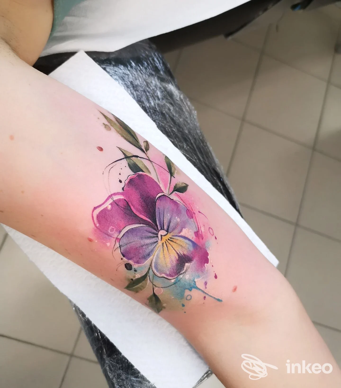 Tatuaggio in stile acquerello su pelle, raffigurante una viola colorata su un avambraccio, con sfumature rosa, viola e giallo e dettagli verdi.