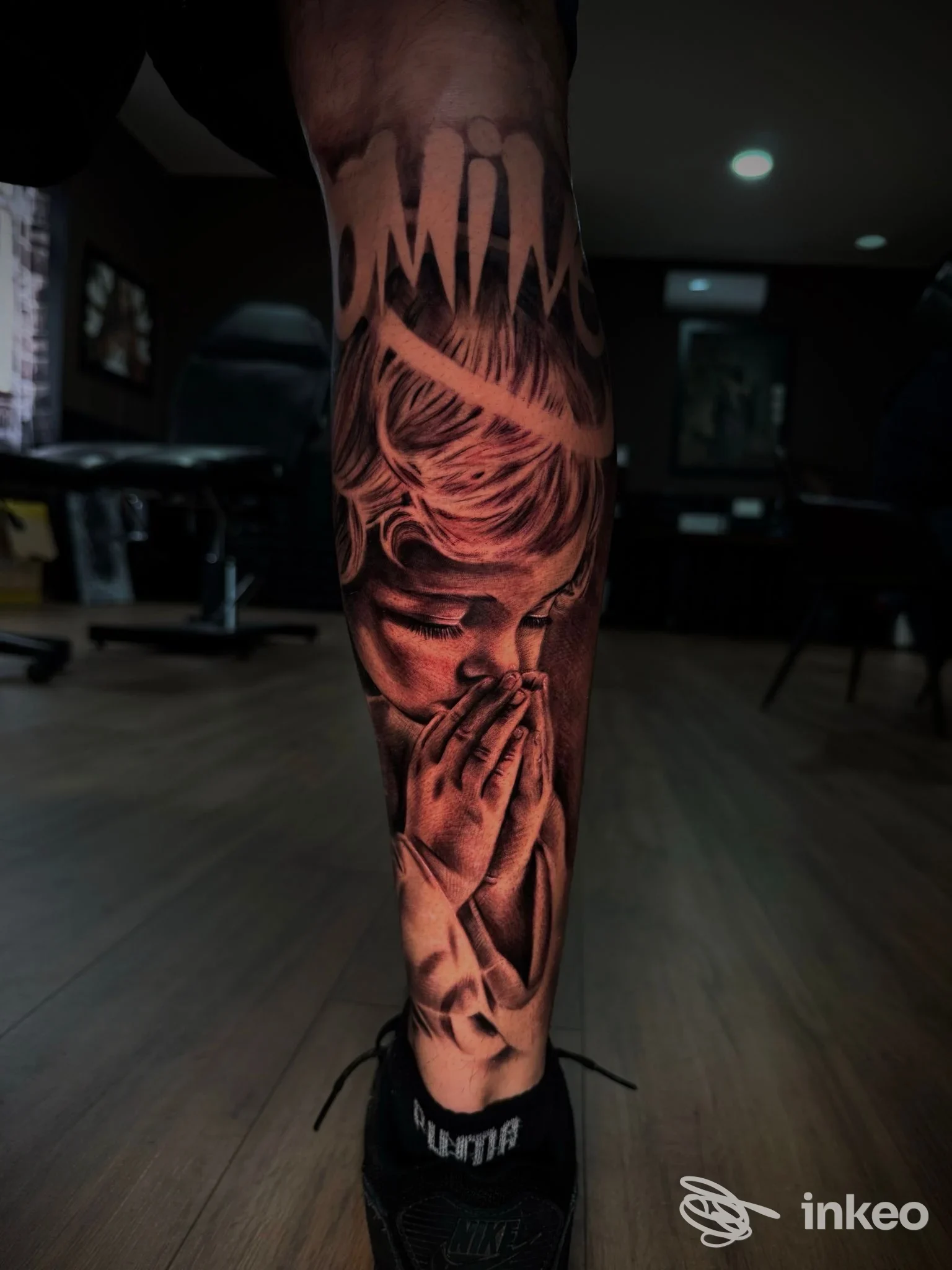 Tatuaggio realistico sulla gamba raffigurante mani in preghiera, realizzato sulla pelle; luce soffusa evidenzia i dettagli.