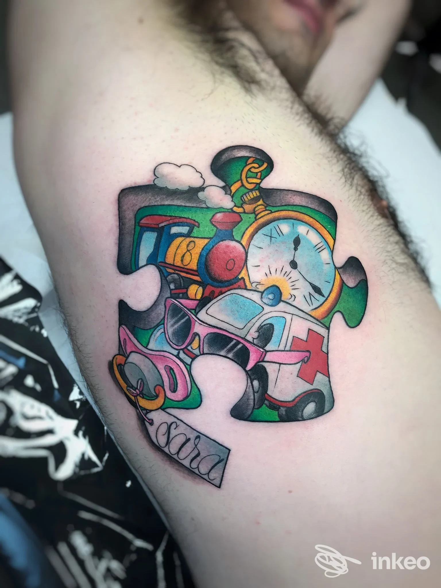 Tattoo colorato a tema puzzle su braccio, raffigurante trenino, orologio, auto e nuvole in stile vivace, con bordi neri, nessun volto visibile.