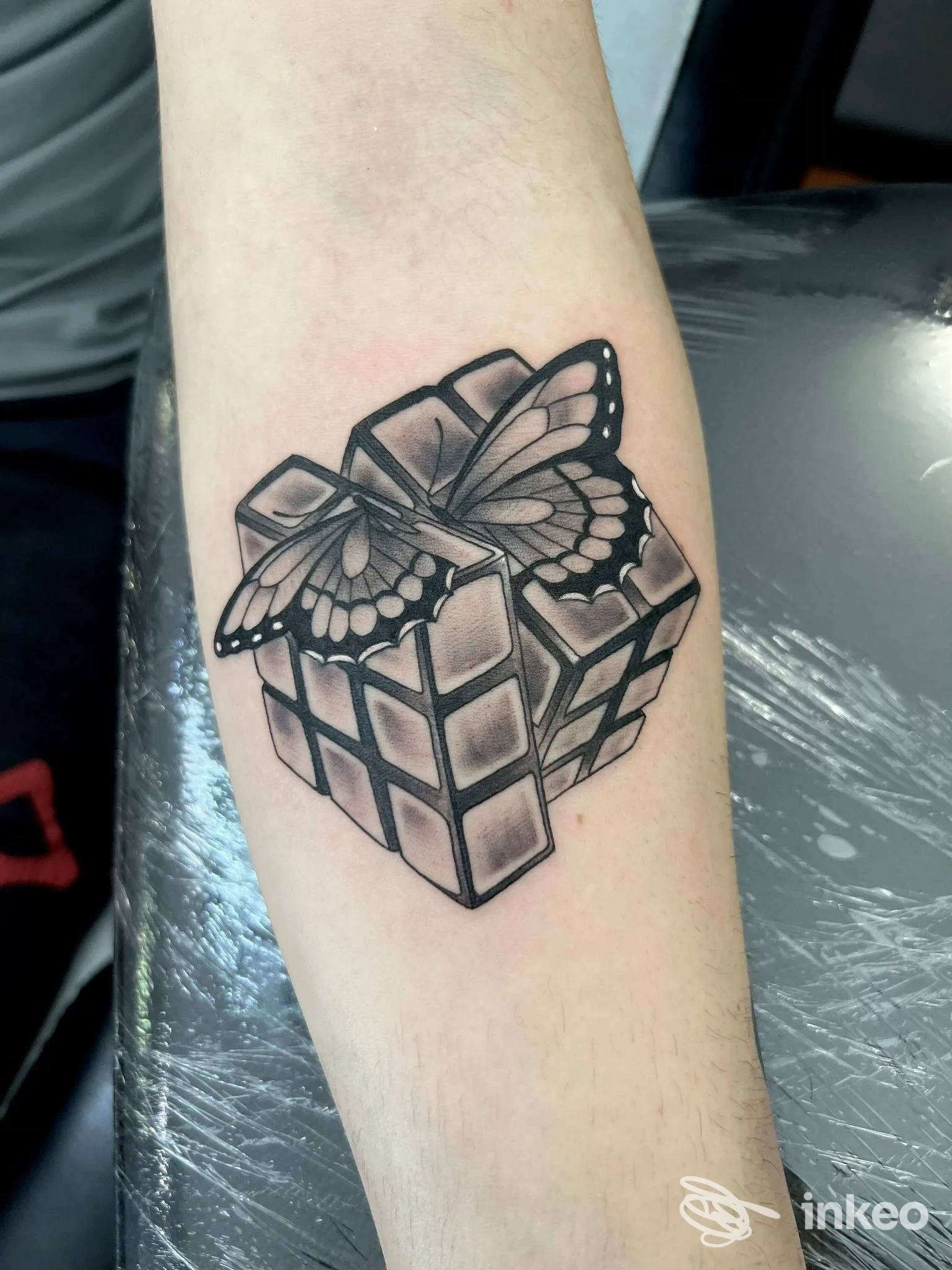 Tatuaggio sull'avambraccio raffigurante una farfalla che emerge da un cubo, realizzato in stile linework nero.