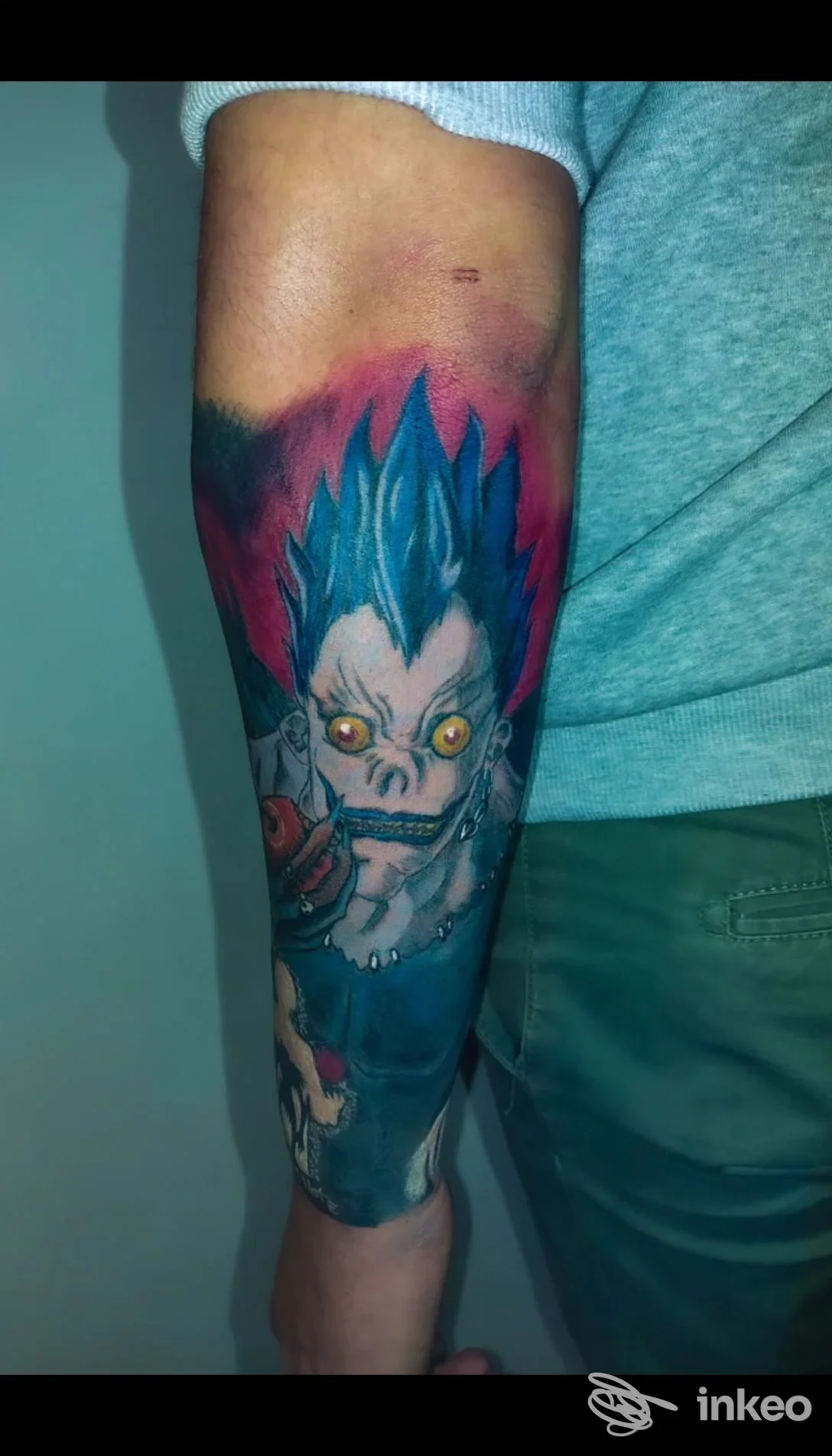 Tatuaggio colorato su avambraccio raffigurante un personaggio fantasy/anime con capelli azzurri, occhi luminosi, oltre a sfumato di colore nello sfondo.