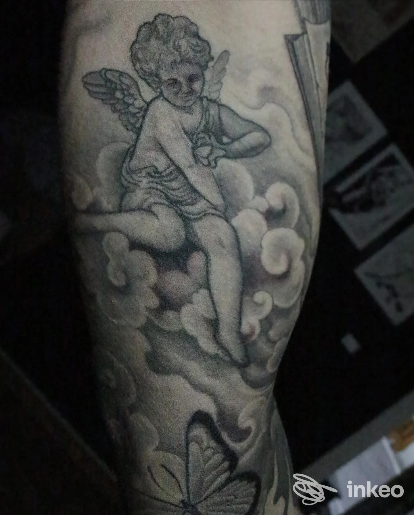 Tatuaggio in bianco e nero di un cherubino con ali su nuvole, situato sull’avambraccio.