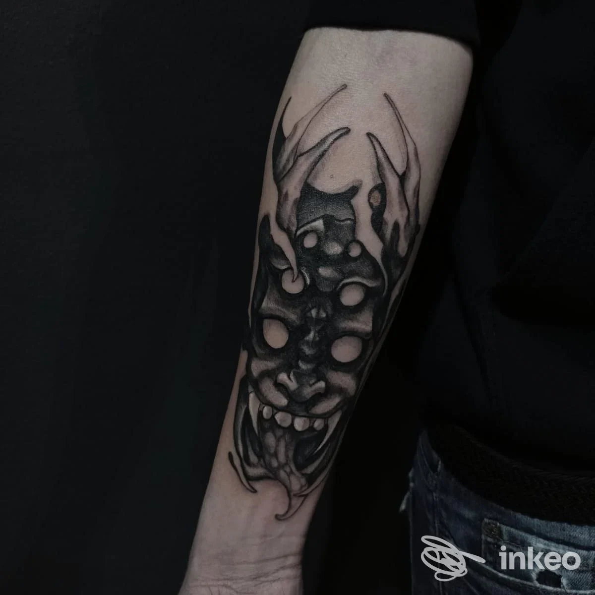 Tatuaggio in bianco e nero sull'avambraccio; figura demoniaca con corna e denti, ombreggiata, su pelle reale.