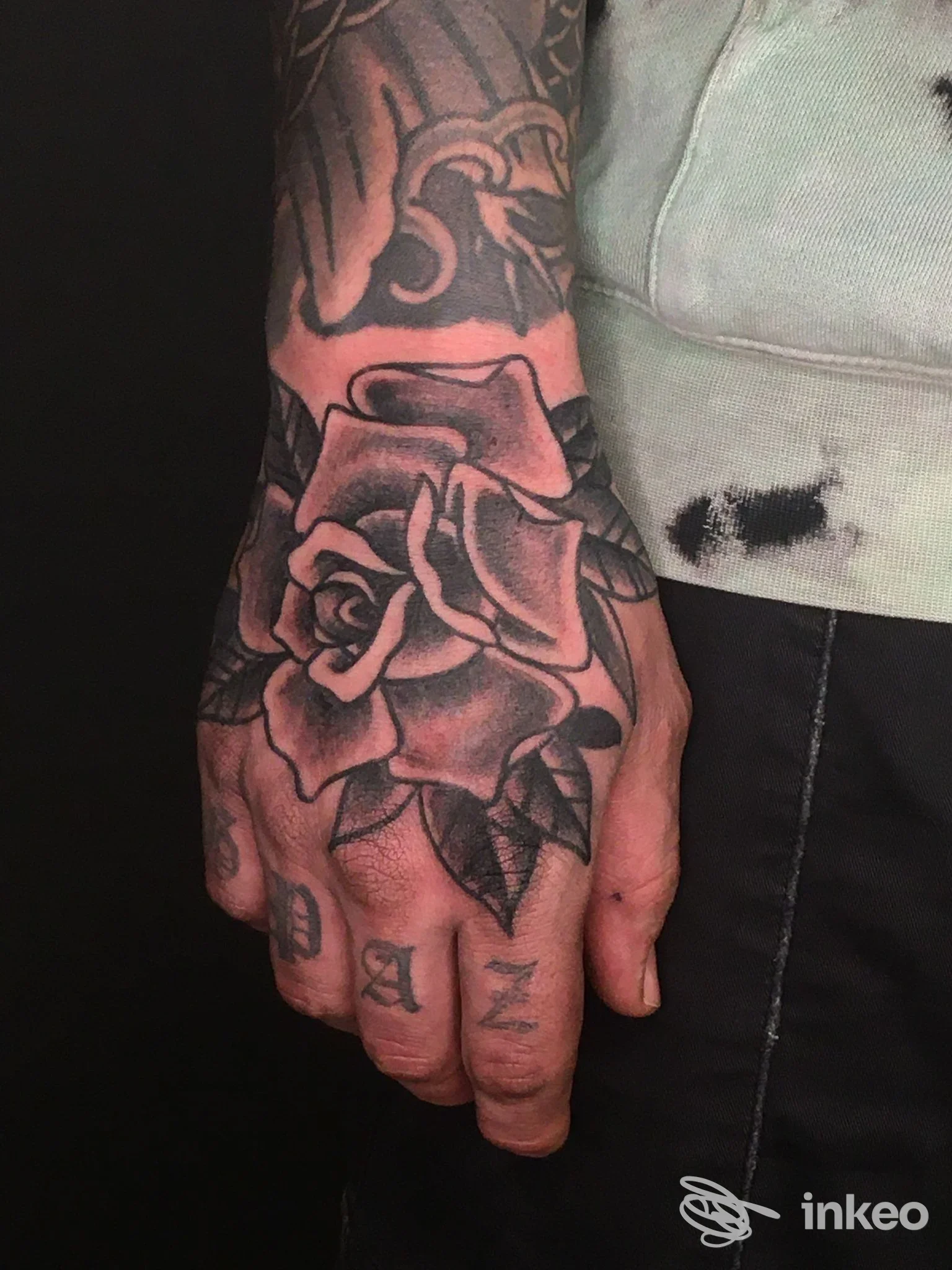 Braccio sinistro visibile con un tatuaggio di rosa grande sull’avambraccio e sulla mano, rilievo marcato e linee scure su pelle.