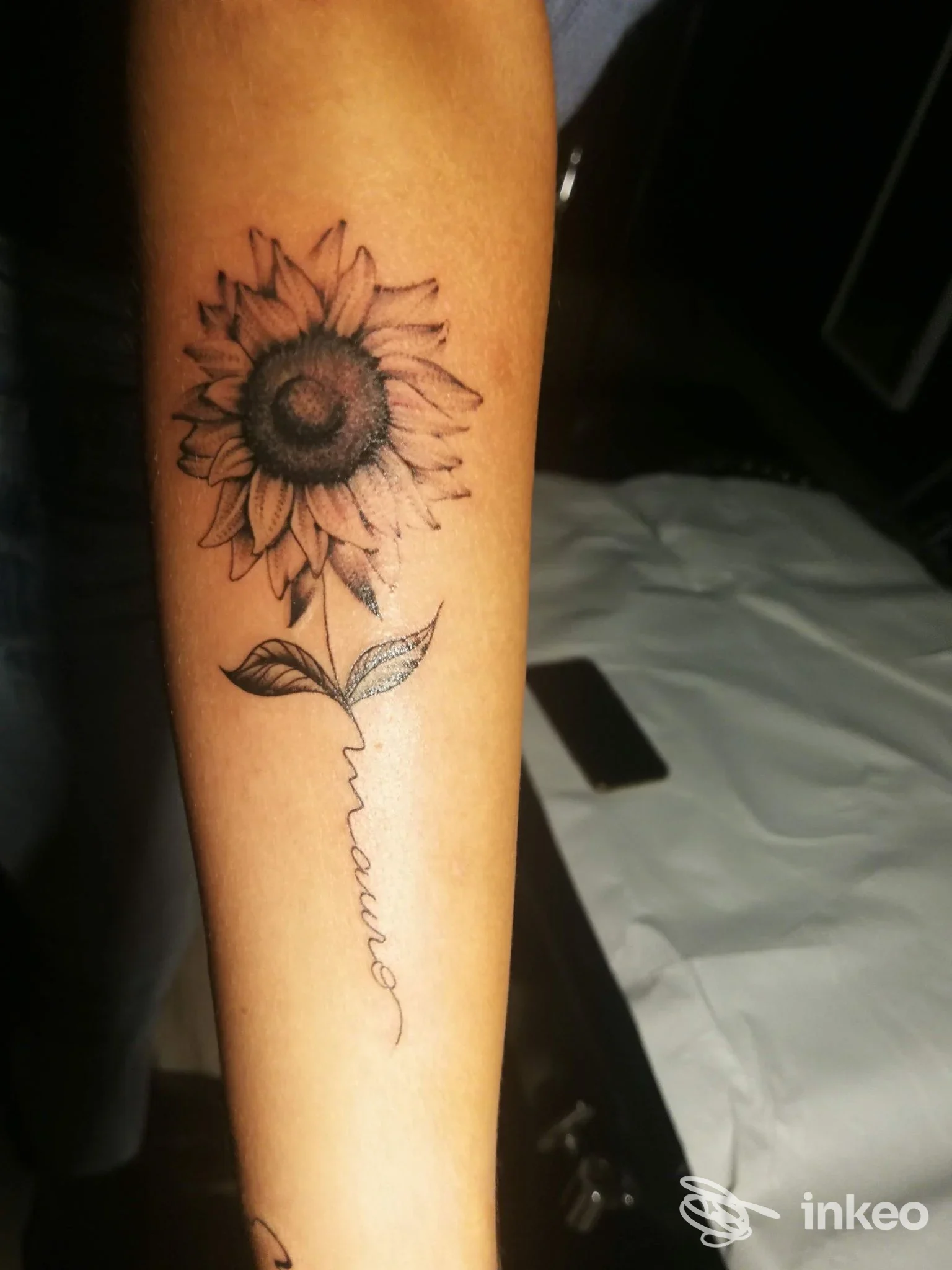 Tatuaggio su pelle raffigurante un girasole disegnato in bianco e nero sull’avambraccio sinistro, con gambo e foglie visibili; stile lineare e dettagliato.