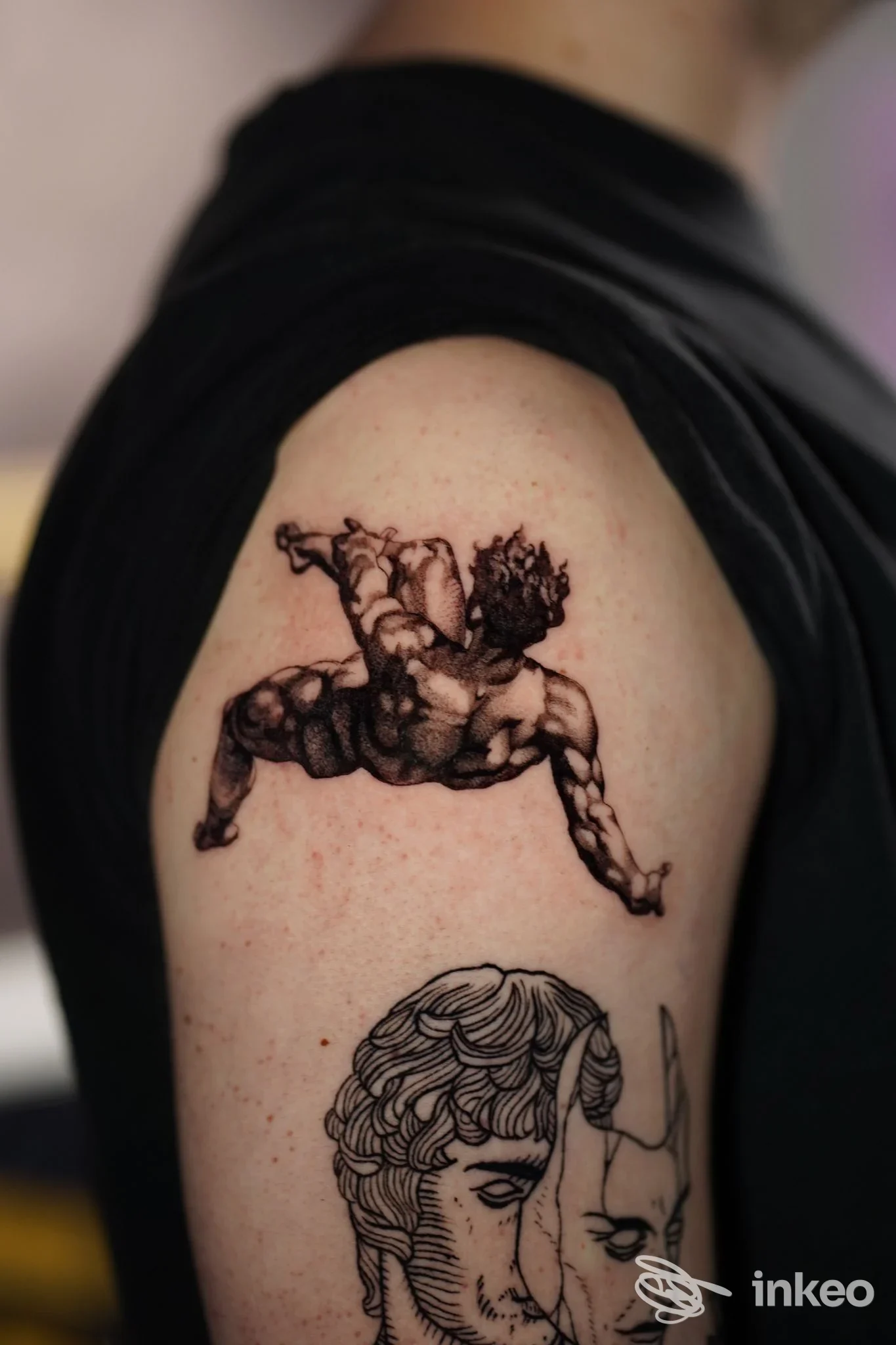Tattoo su braccio con disegno scuro, mostra una figura fantastica o cornice, con una seconda immagine più in basso; pelle esposta senza elementi digitali.
