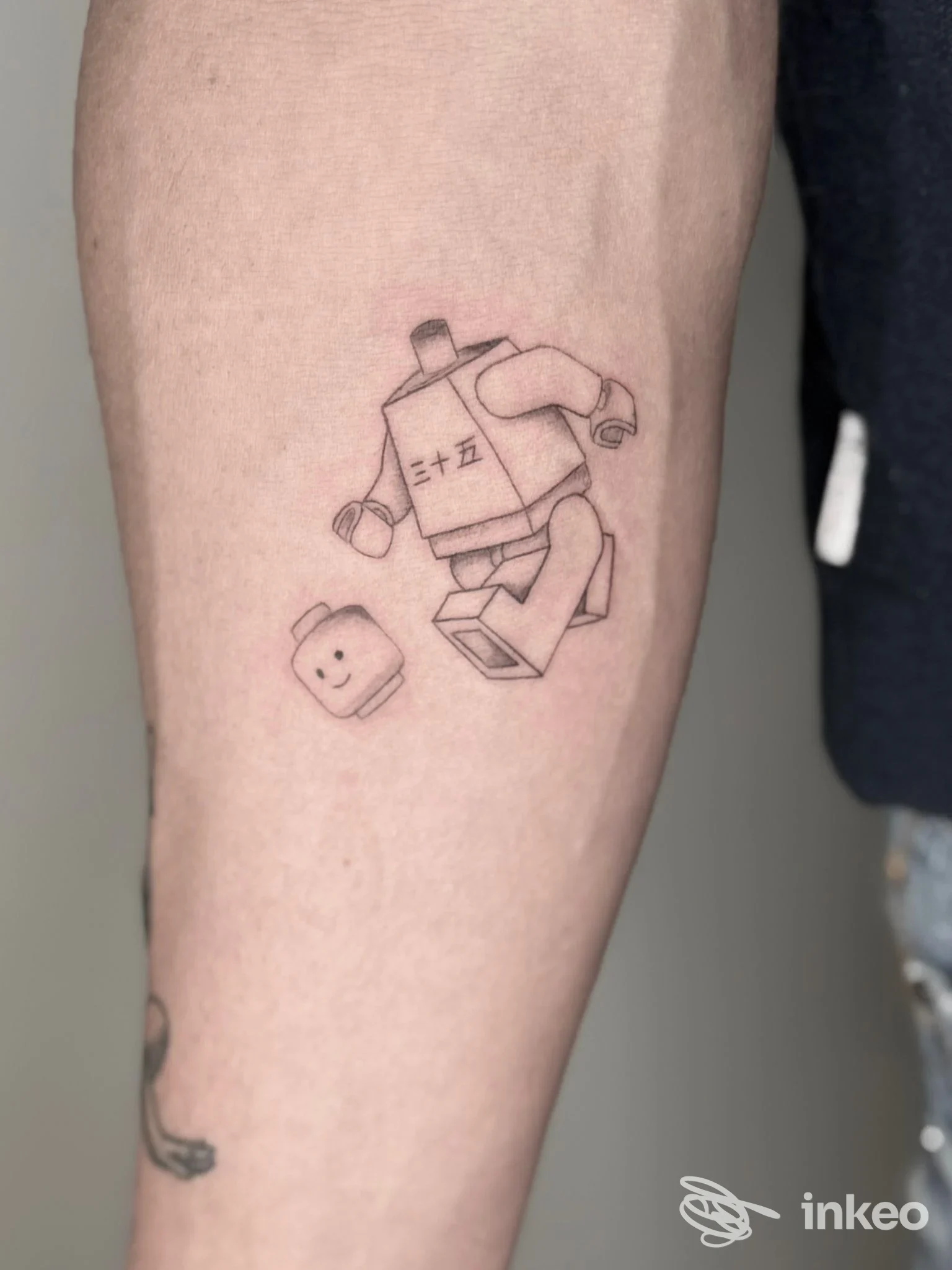 Tattoo su avambraccio raffigurante un piccolo robot stilizzato con un personaggio accanto, visibile chiaramente sulla pelle.