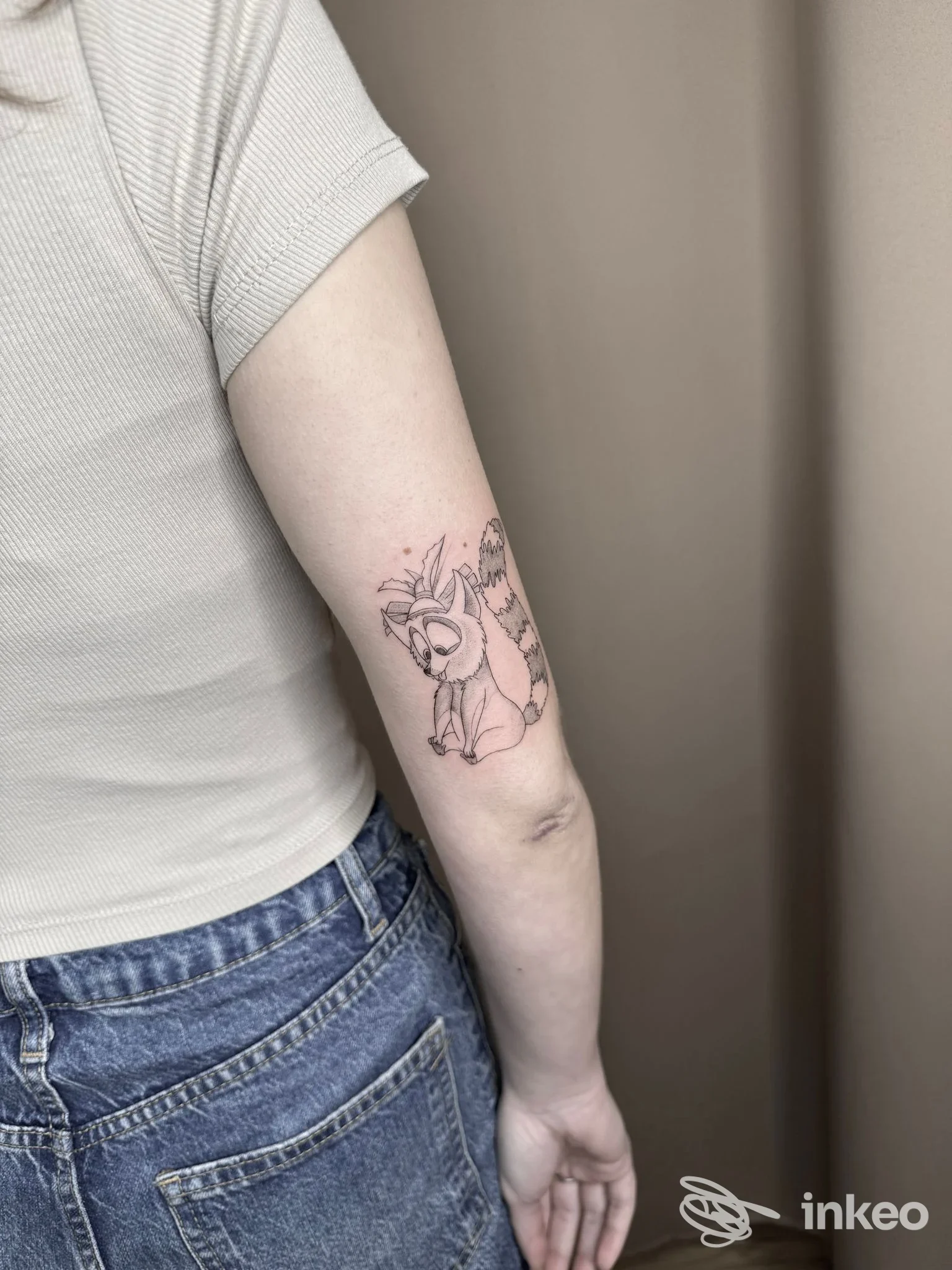 Braccio di una persona con tattoo di un animale stilizzato visibile chiaramente, vestendo una maglietta corta color champagne e jeans, sfondo neutro.