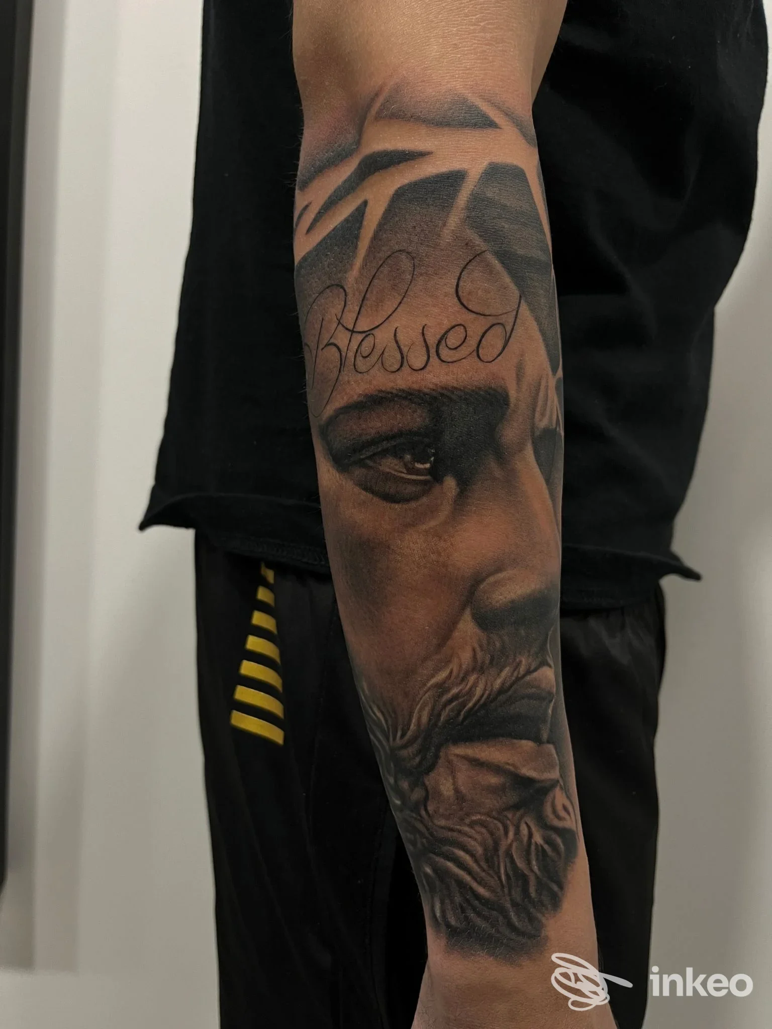 Avambraccio sinistro di persona, tatoo realistico in bianco e nero con ritratto e scritta Blessed sull'avambraccio, sfondo scuro e abbigliamento casual.