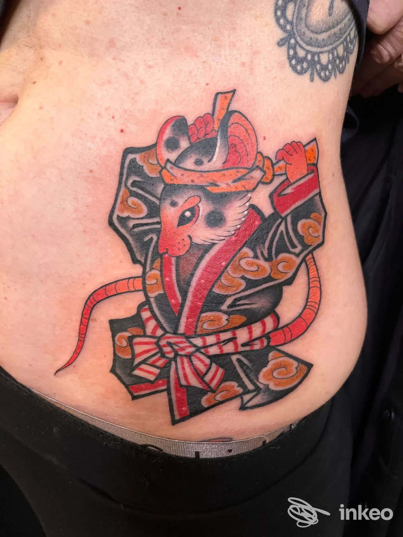 Tattoo sull’addome raffigurante un topo stilizzato con motivi ornamentali in stile giapponese, colori rosso/nero/grigio e nastri decorativi.