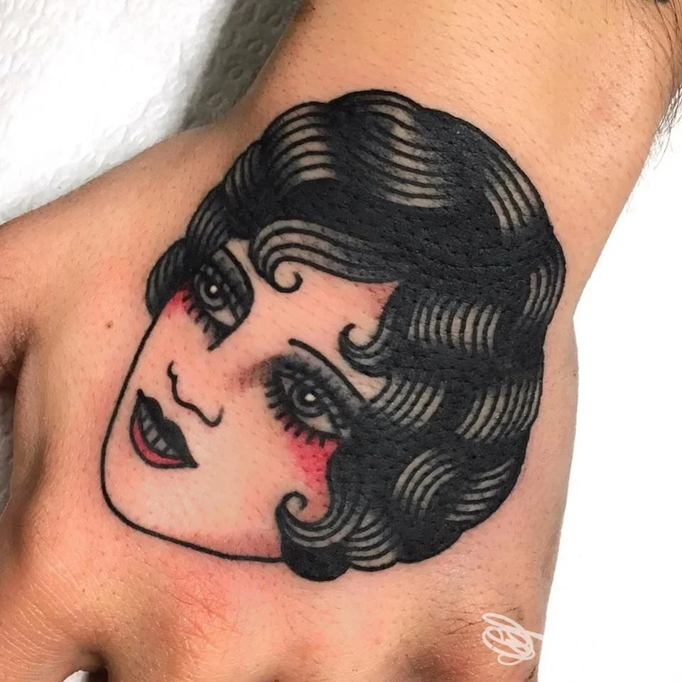 Tattoo sul dorso della mano raffigurante una faccia femminile in stile cartoonesco su pelle, realizzato in inchiostro nero con accenti rossi sulle guance.