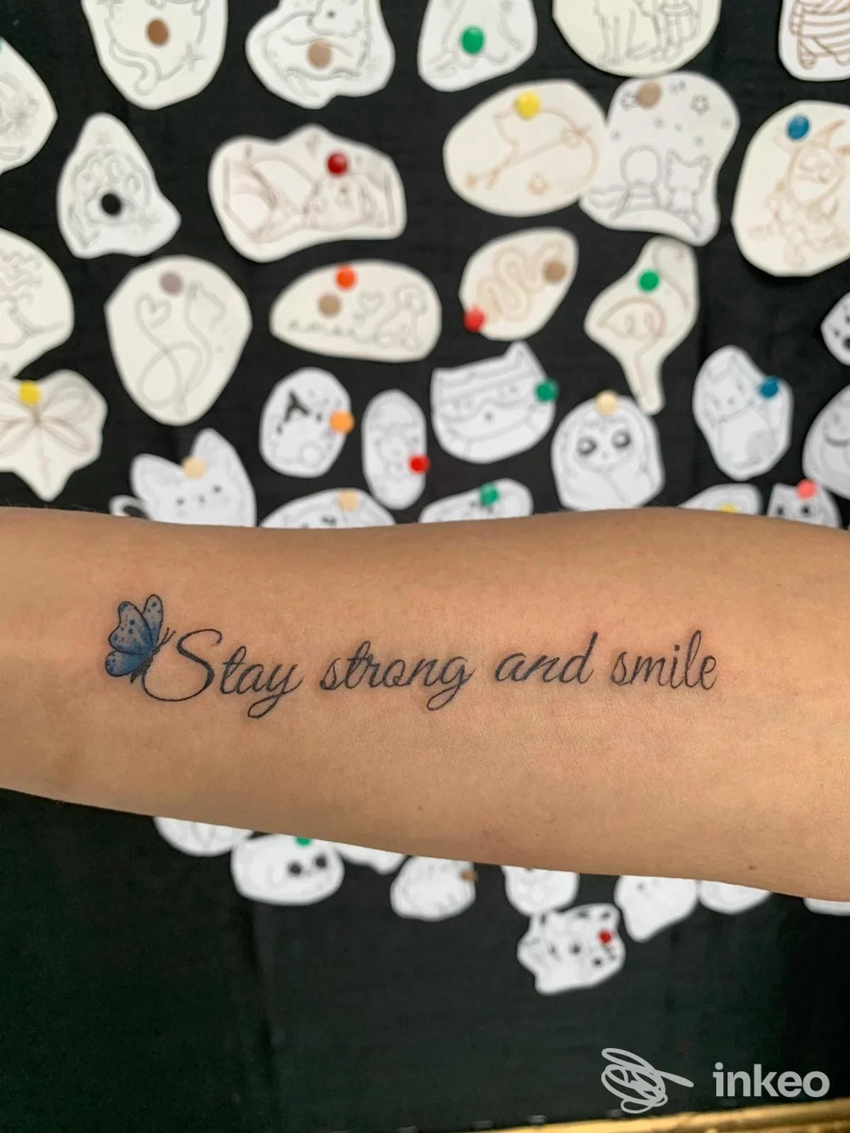 Tattoo sull'avambraccio con la frase Stay strong and smile e una piccola farfalla blu, realizzato direttamente sulla pelle.