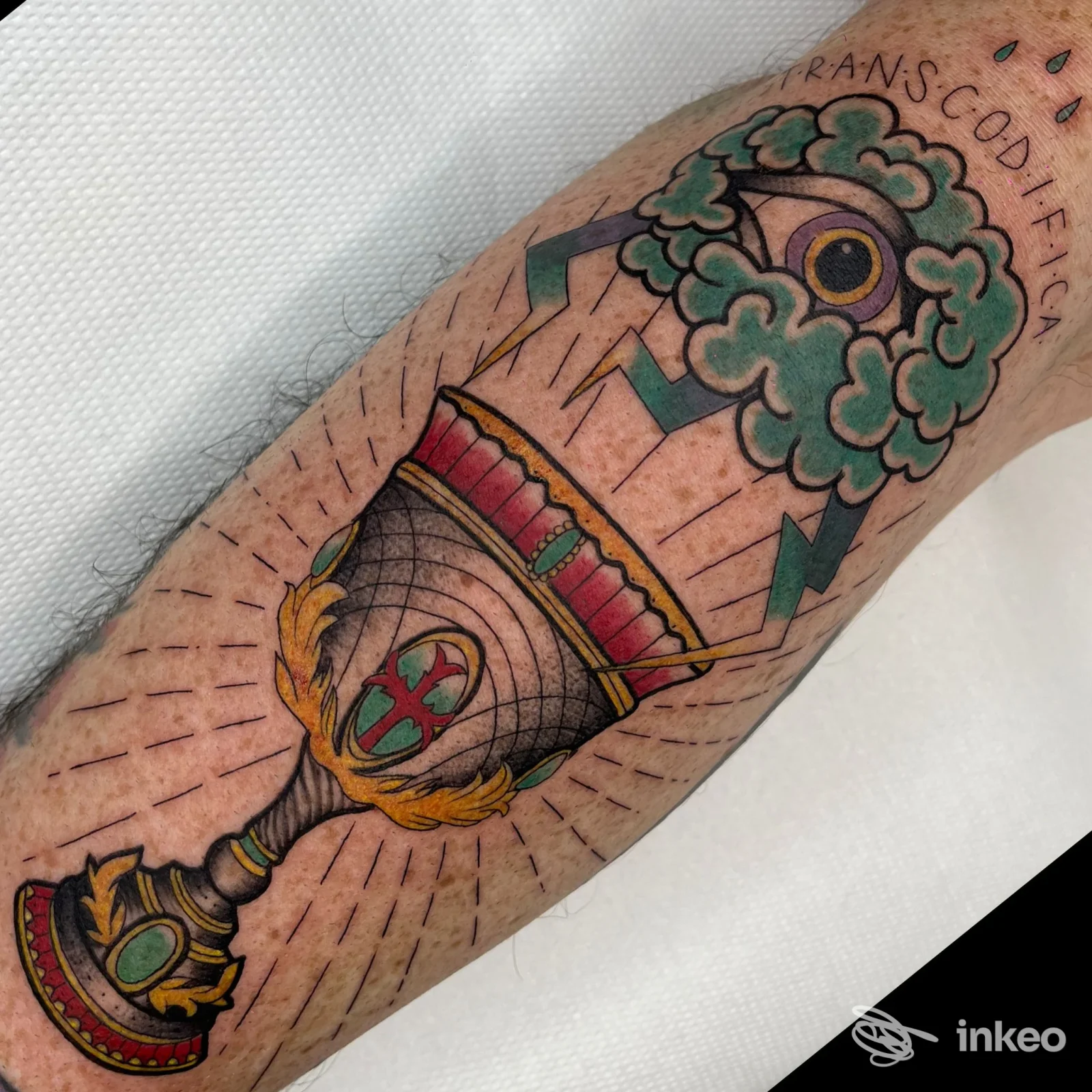 Tattoo a colori sull’avambraccio raffigurante un calice decorato con corona e un motivo di nuvole verdi e occhio, sfondo chiaro in studio.