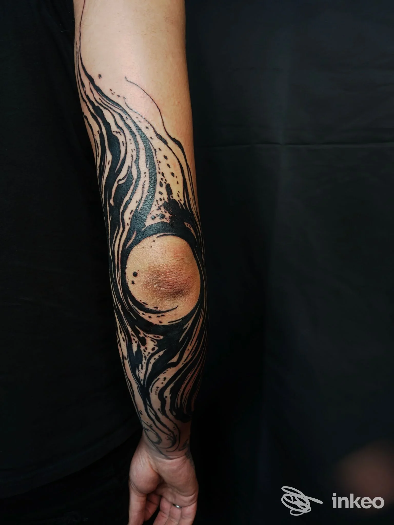 Tatuaggio astratto in nero sull’avambraccio sinistro, contorno curvo con linee e schizzi scuri, sfondo scuro, nessun volto visibile.