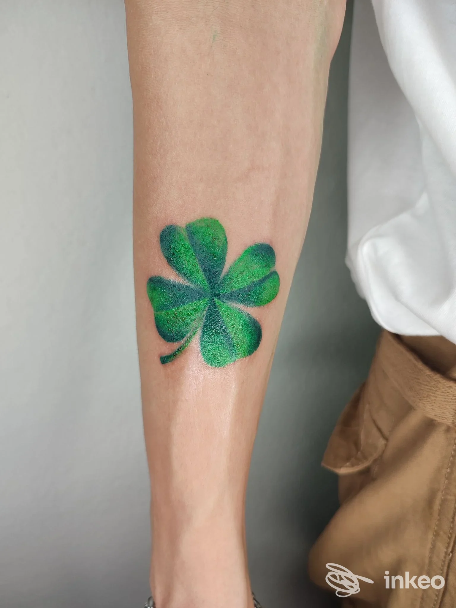 Dettaglio di un tatuaggio verde a forma di quadrifoglio sull'avambraccio di una persona, sfondo neutro.